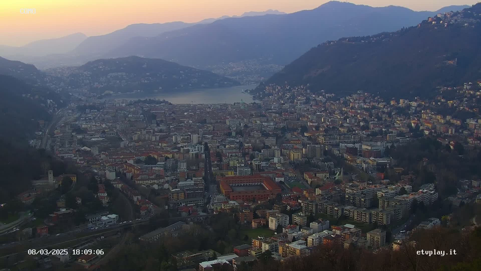 A sprawling city nestled in a valley between mountains, overlooking a lake, under a dusky, sunset sky.