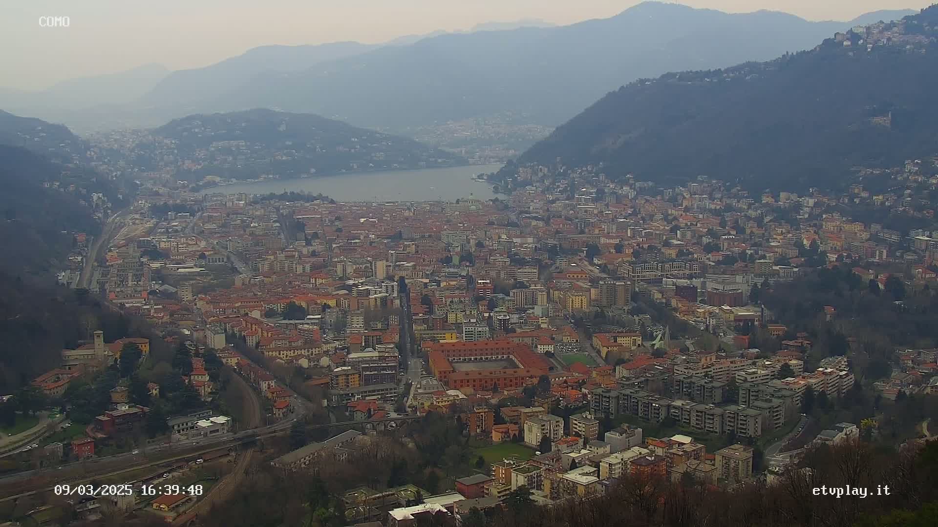 A hazy, high-angle view shows a sprawling city nestled between mountains and a lake under an overcast sky.