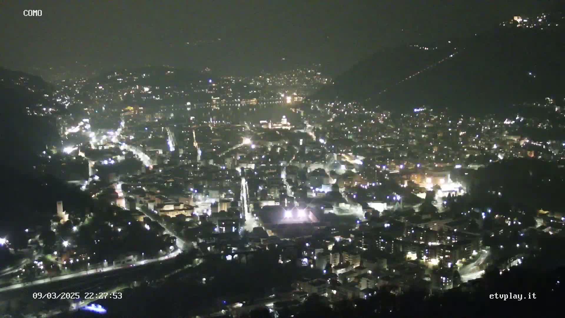 A nighttime, high-angle view of a city illuminated by streetlights and building lights, nestled within hills under a dark, clear sky.