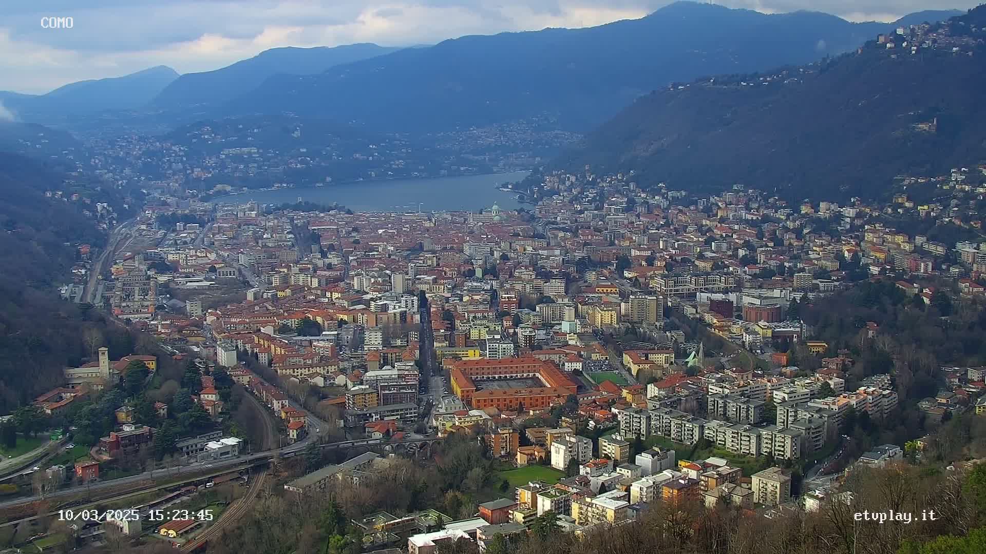 A sprawling city nestled in a valley between mountains, overlooking a lake under a mostly cloudy sky.