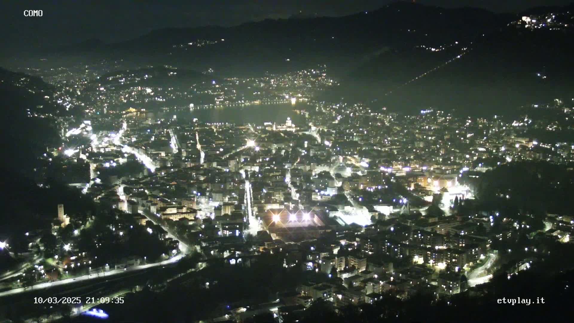 A nighttime, aerial view of a city illuminated by streetlights, nestled in a valley near a body of water under a clear, dark sky.