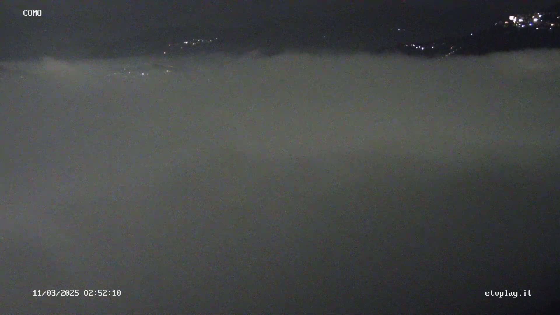 A nighttime scene of a thick layer of fog covering a landscape, with faintly visible lights in the distance.