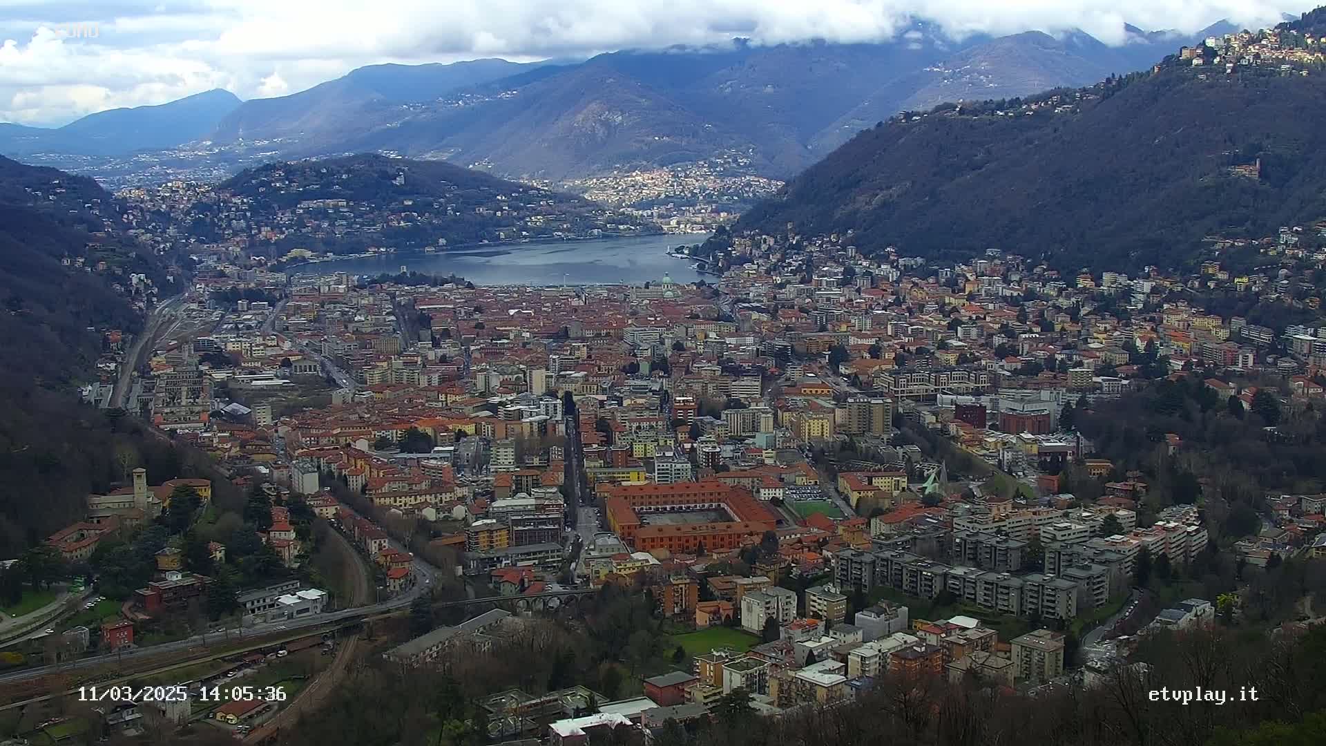 A sprawling city nestled in a valley between mountains, under a partly cloudy sky, with a body of water visible in the distance.