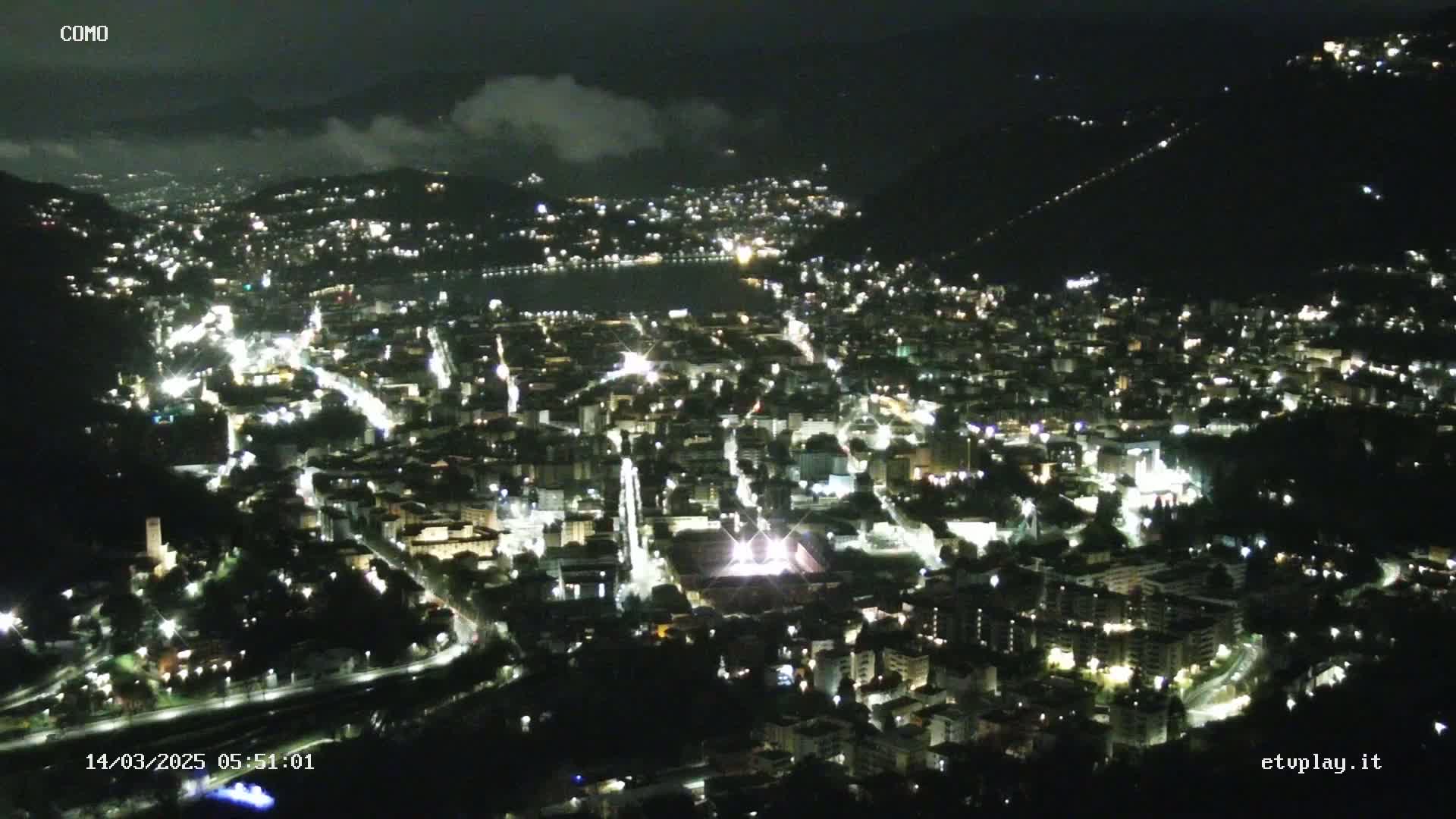 A nighttime, high-angle view of a city illuminated by lights, nestled in a valley with a body of water visible in the distance, under a partly cloudy sky.