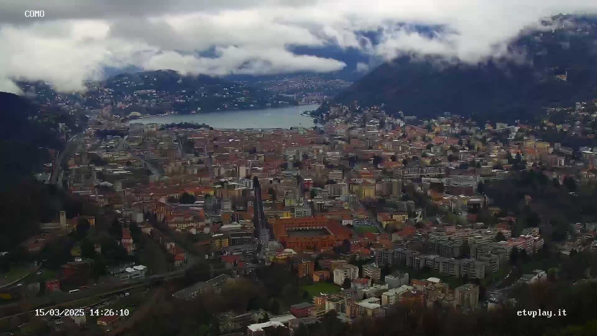 A sprawling city nestled between a lake and mountains under a cloudy sky.