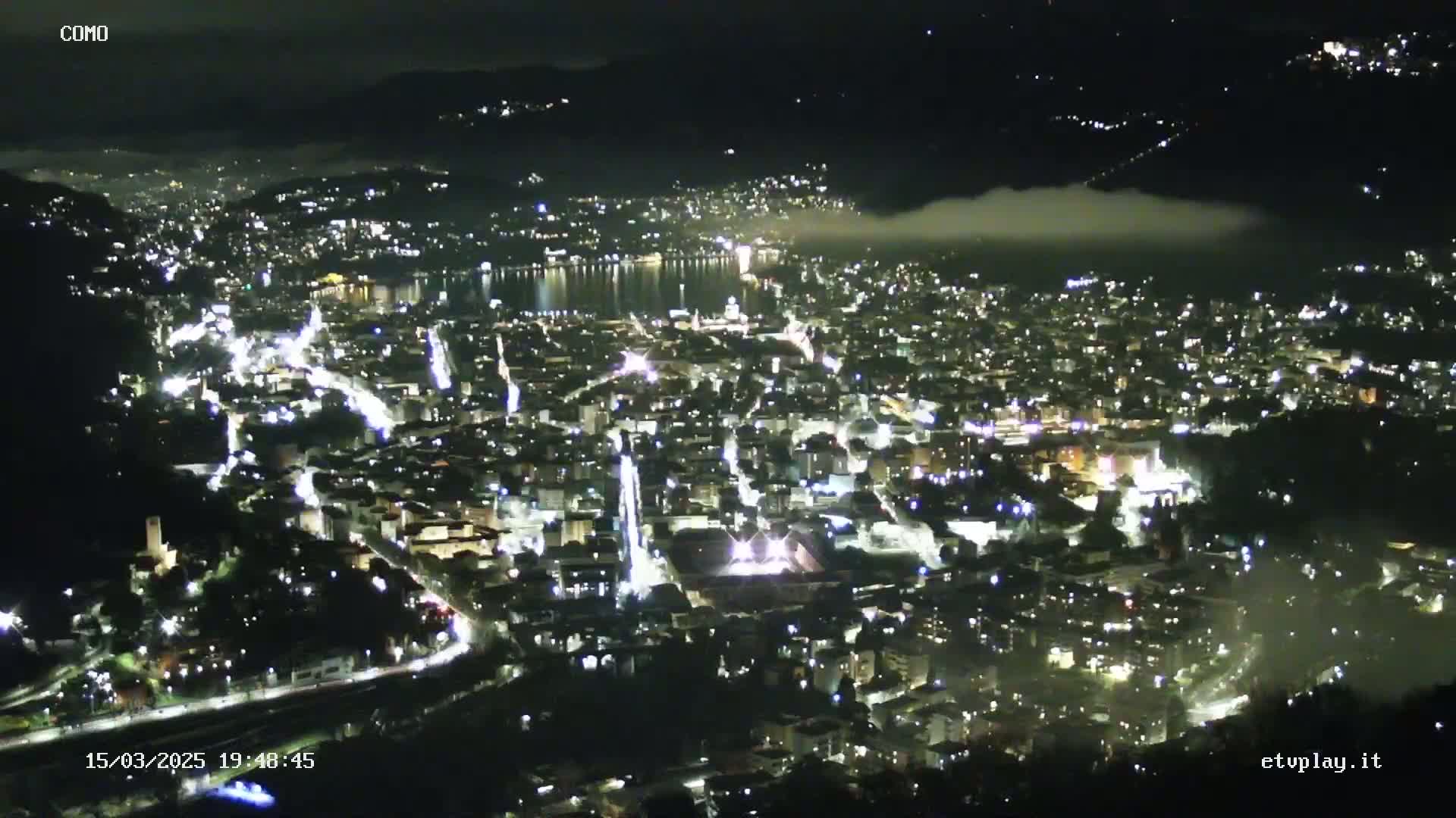 A nighttime, high-angle view of a city illuminated by lights, with fog partially obscuring a body of water in the distance.