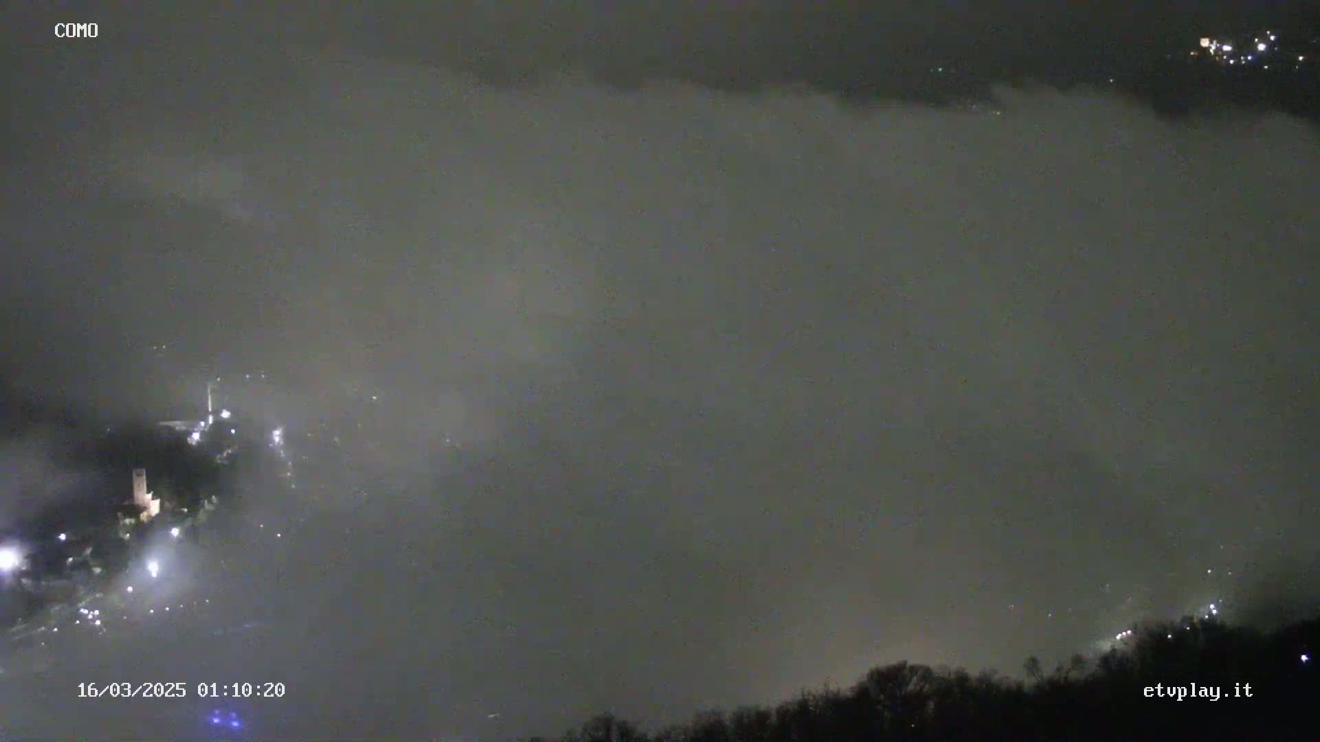 A nighttime, foggy landscape shows faintly lit buildings and trees below a thick layer of fog.