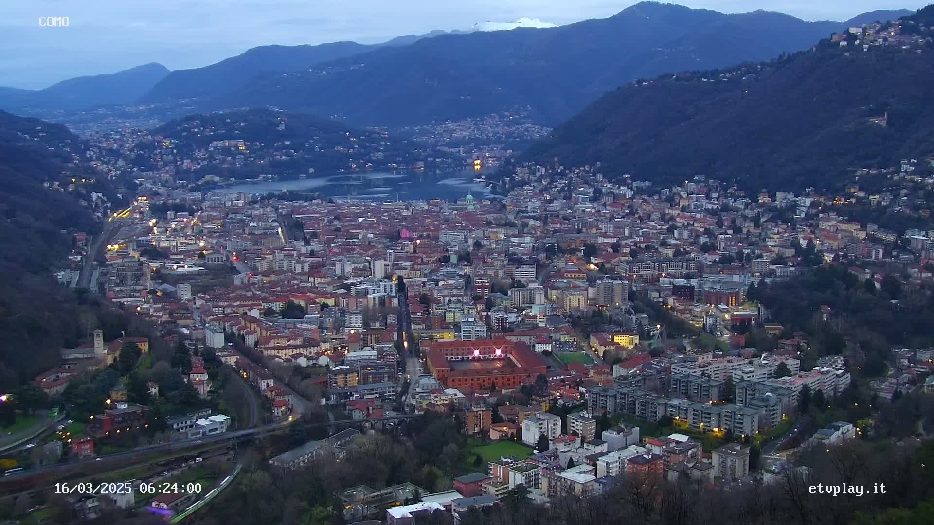 A nighttime, high-angle view of a city nestled in a valley between mountains, illuminated by streetlights and with a body of water visible in the distance.
