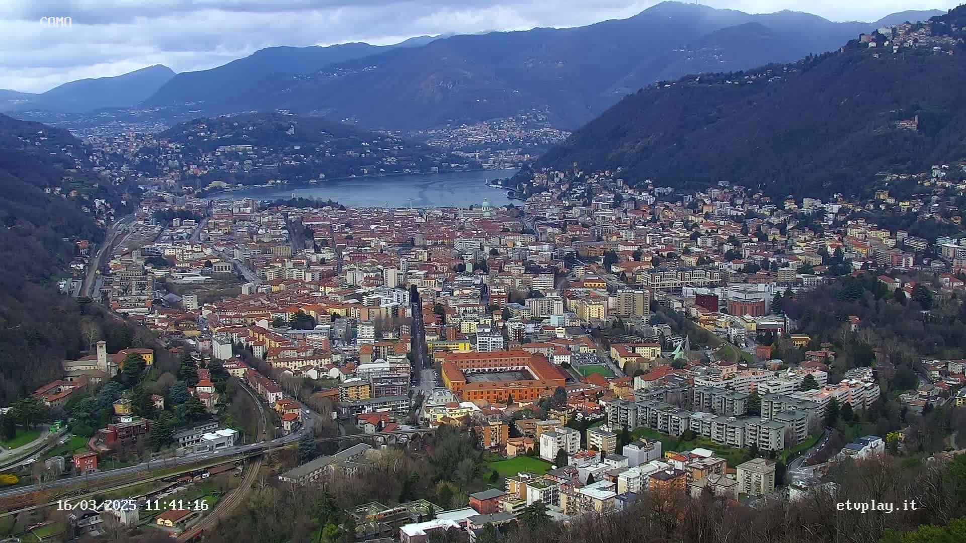 A sprawling city nestled in a valley between mountains, overlooking a lake under an overcast sky.
