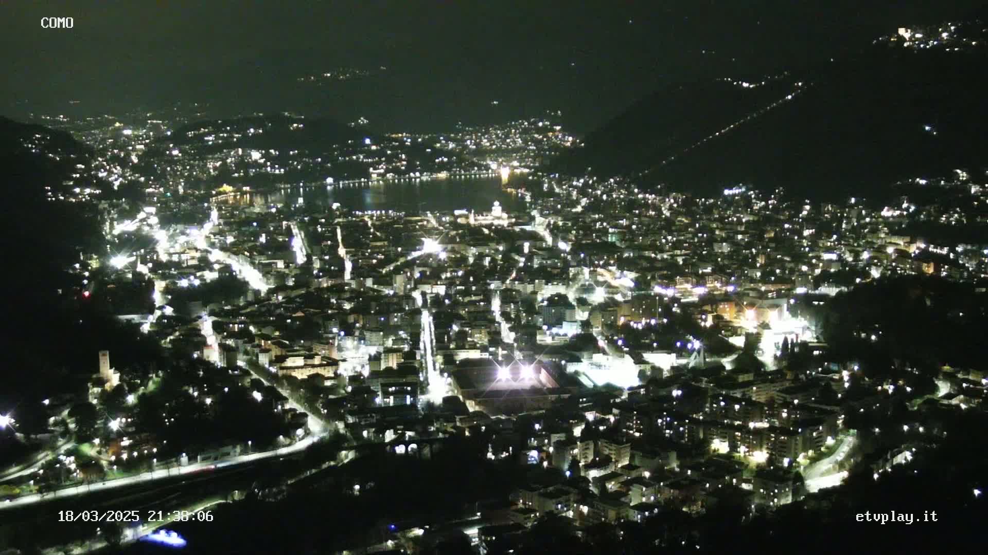 A nighttime aerial view of a city illuminated by streetlights, situated next to a body of water, under a clear, dark sky.