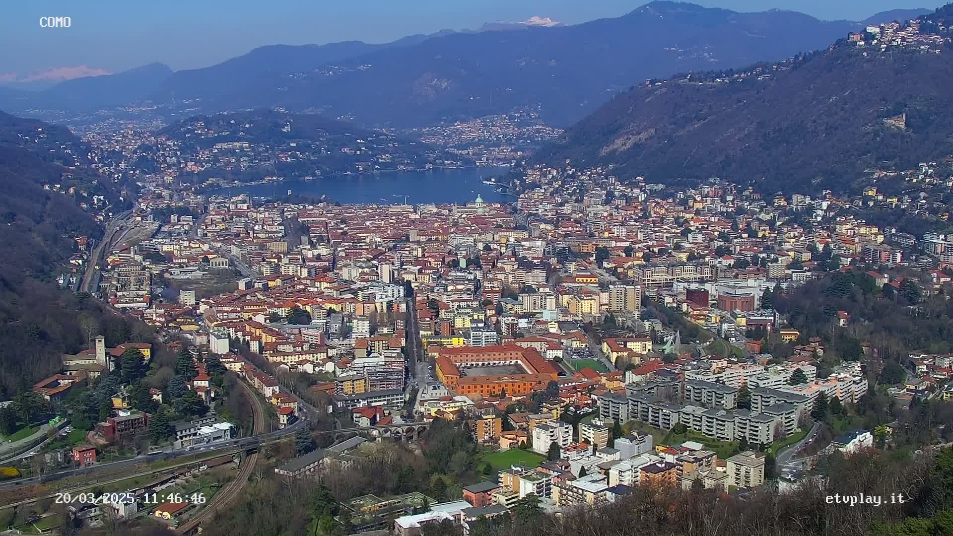 A sprawling city nestled in a valley between mountains, overlooking a lake under a clear, sunny sky.