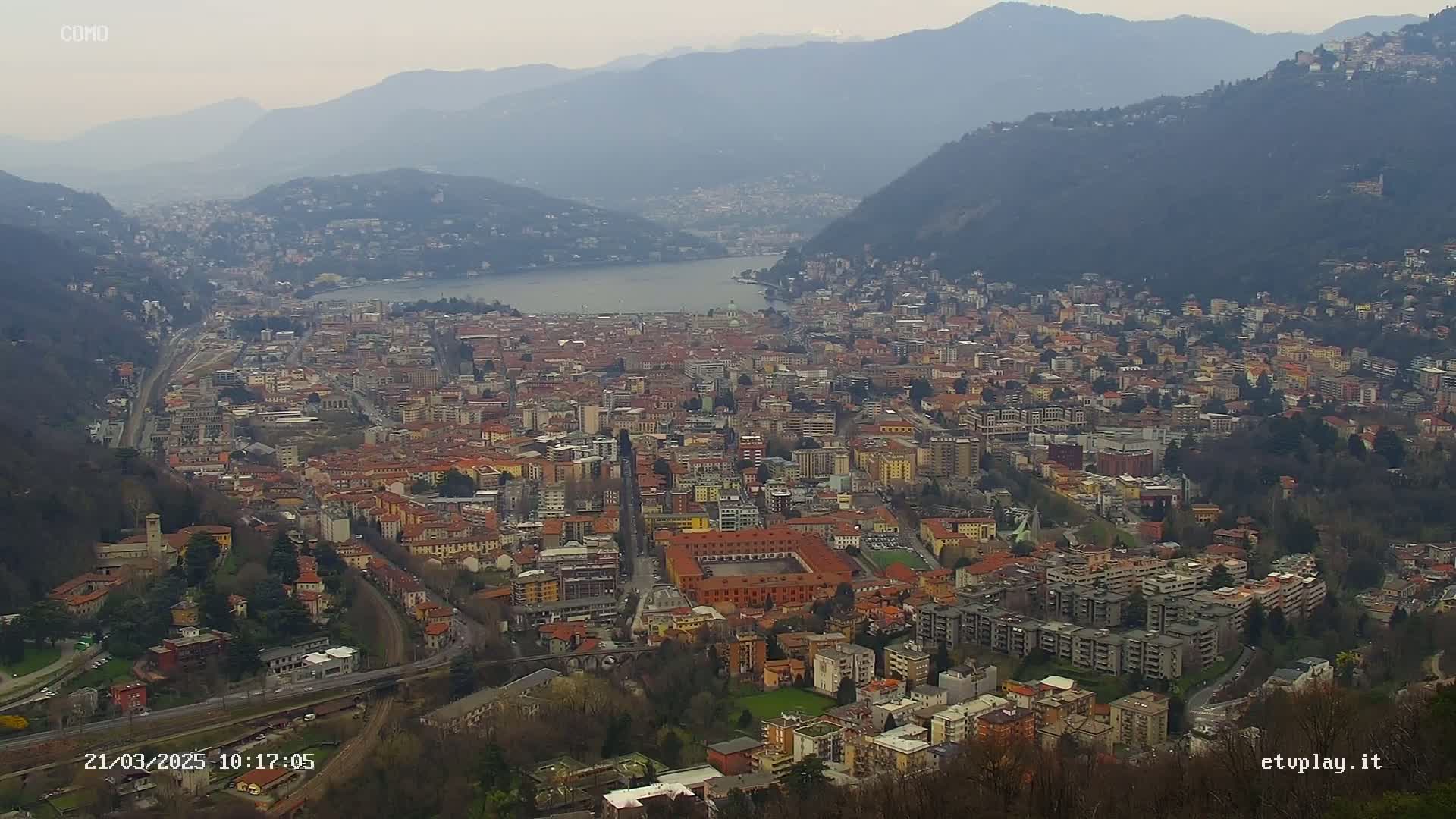 A sprawling city nestled between mountains and a lake under an overcast sky.