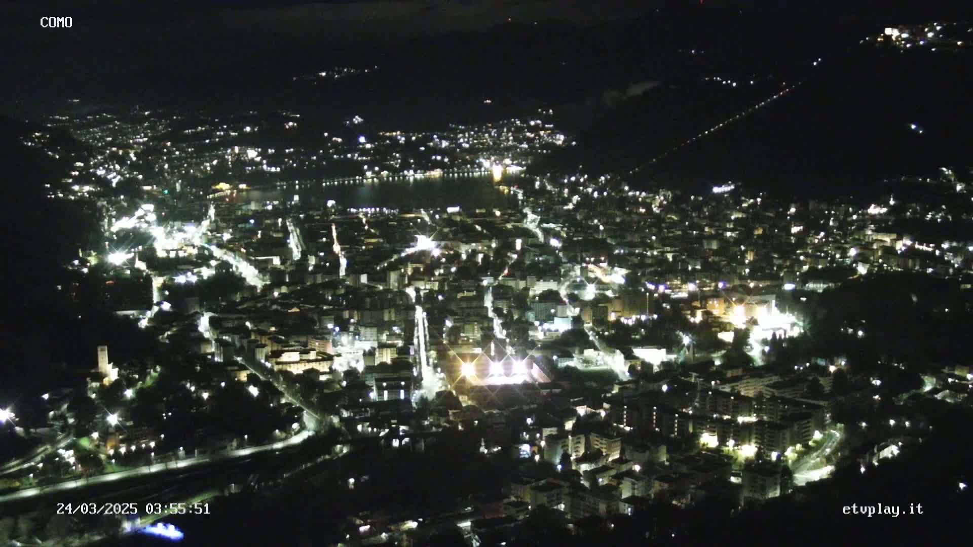A nighttime aerial view of a city illuminated by streetlights, nestled beside a dark body of water under a clear sky.