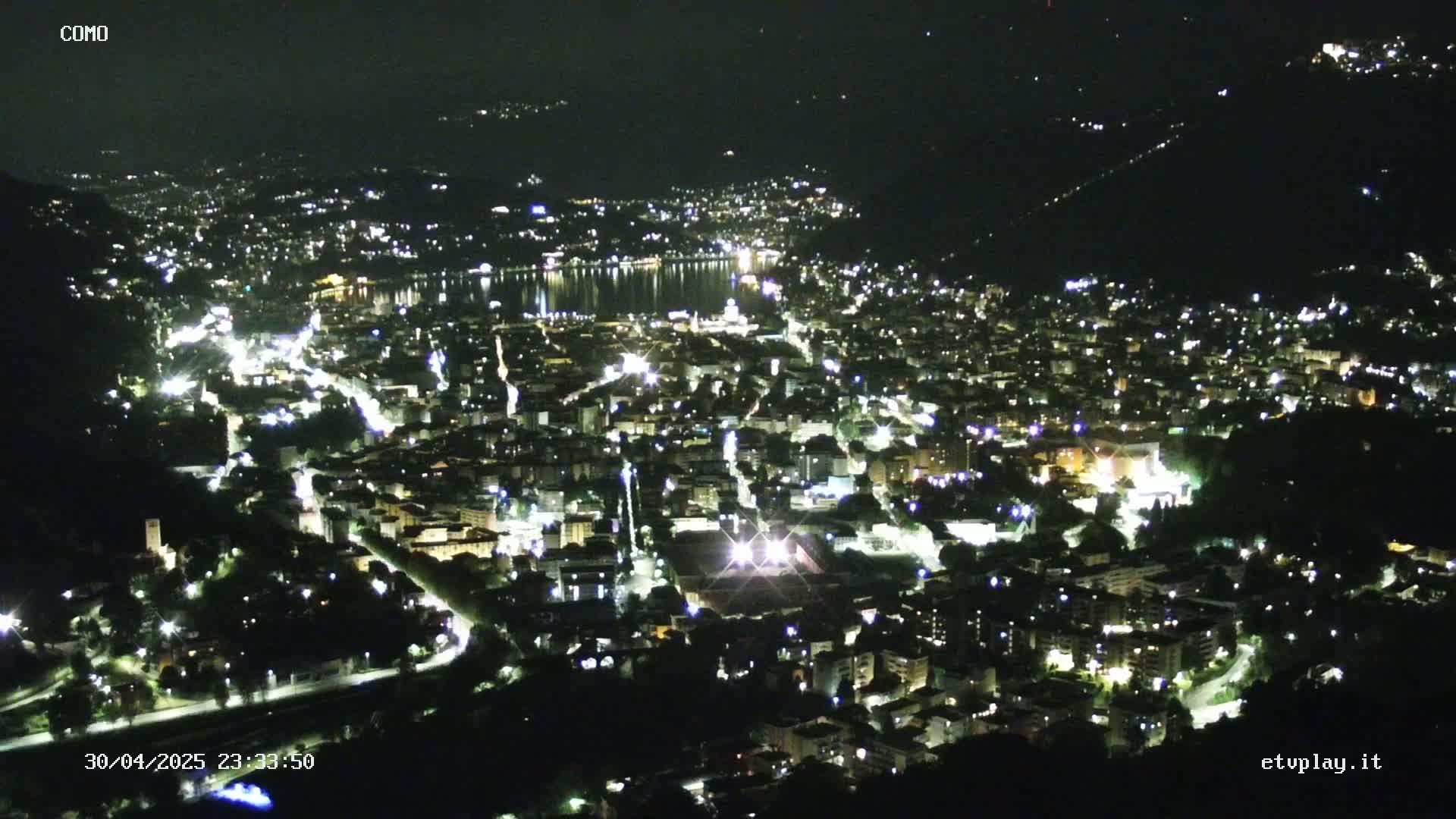 A nighttime aerial view of a city situated near a body of water, with scattered lights illuminating buildings and roads under a clear sky.