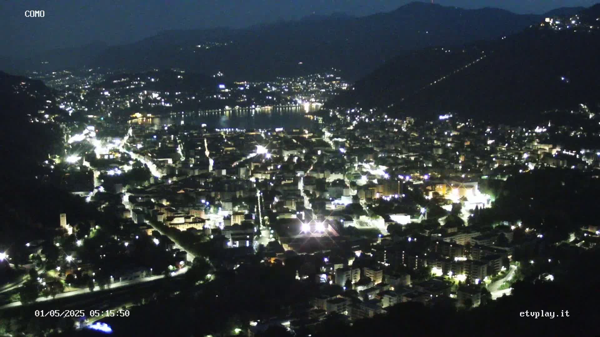 A nighttime, aerial view of a city illuminated by lights, nestled beside a dark body of water, surrounded by hills.