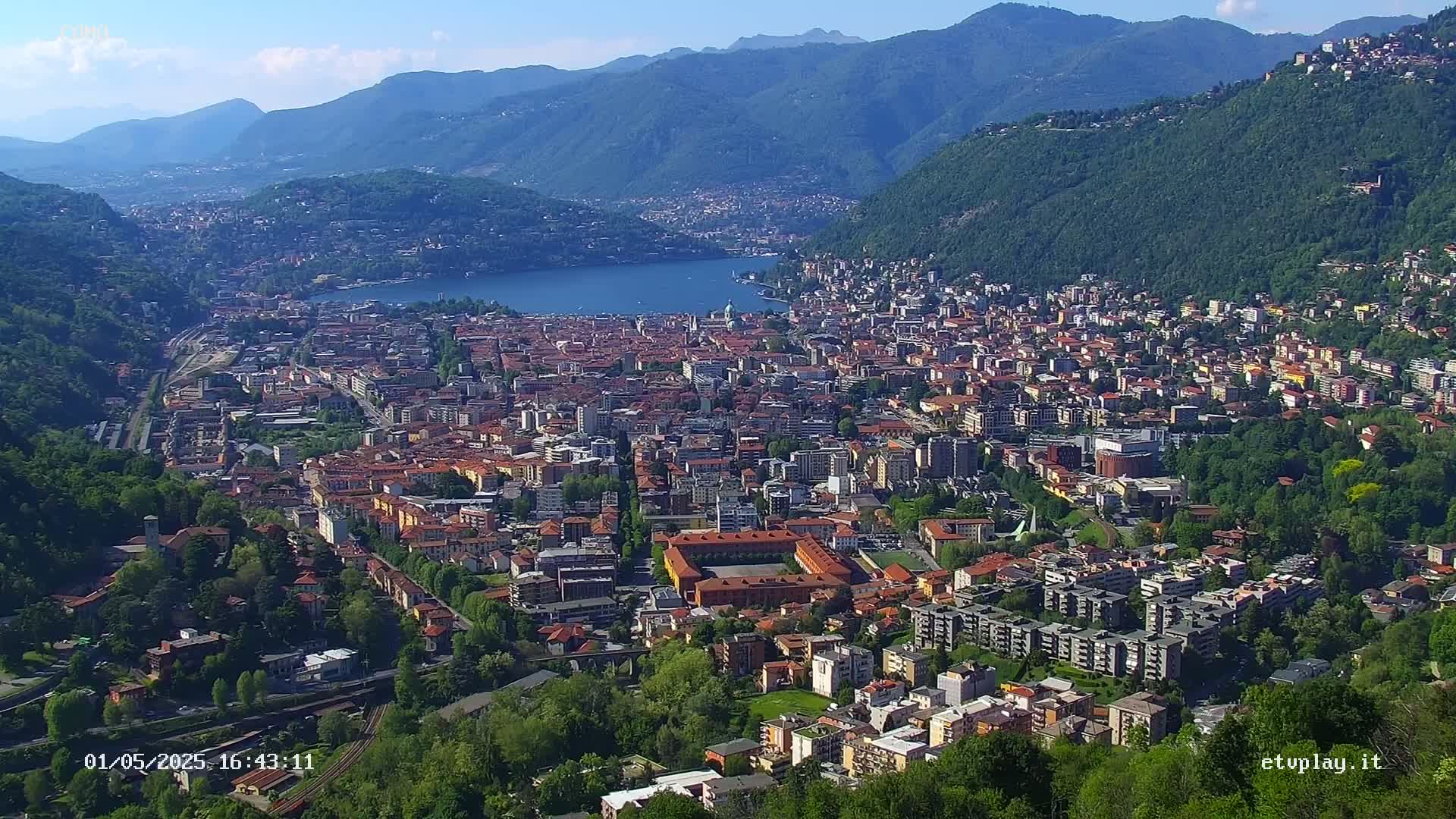 An aerial view shows a city nestled in a valley between mountains, overlooking a lake under a clear, sunny sky.