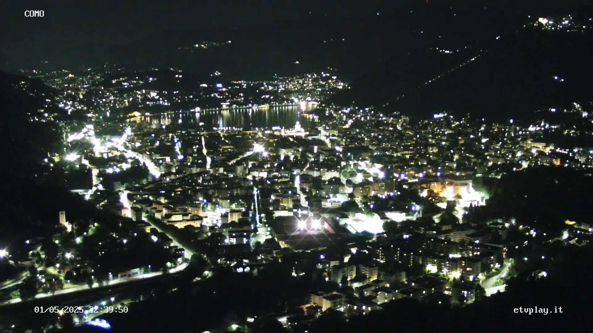 A nighttime aerial view of a city illuminated by lights, situated beside a body of water under a clear, dark sky.