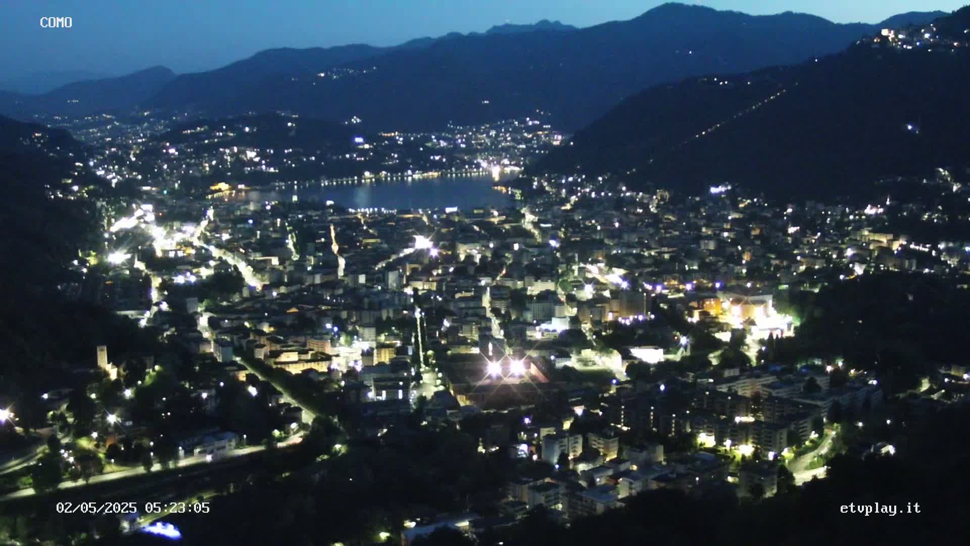 A nighttime aerial view of a city nestled in a valley, illuminated by numerous lights, with a dark body of water visible in the distance under a clear, dark sky.