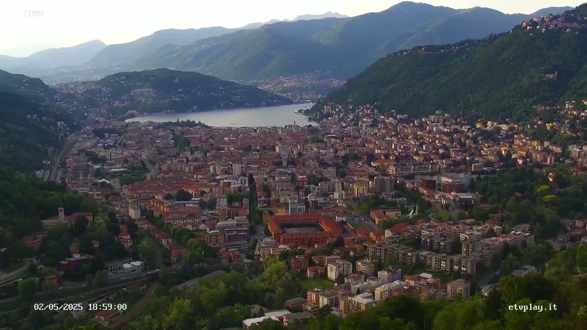 An aerial view shows a city nestled between mountains and a lake under a clear sky.