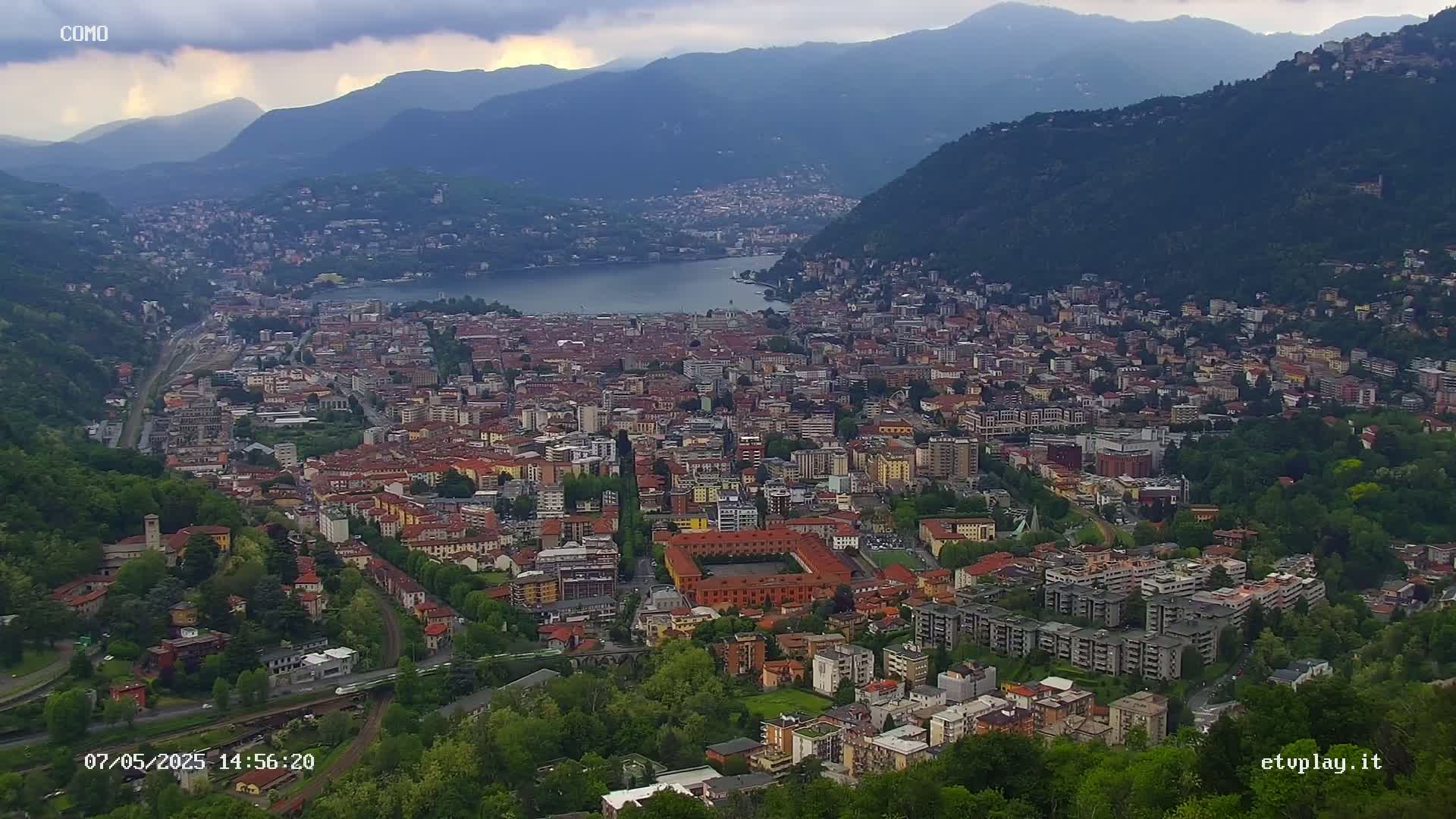 An expansive view of a city nestled between mountains and a lake under a cloudy sky.