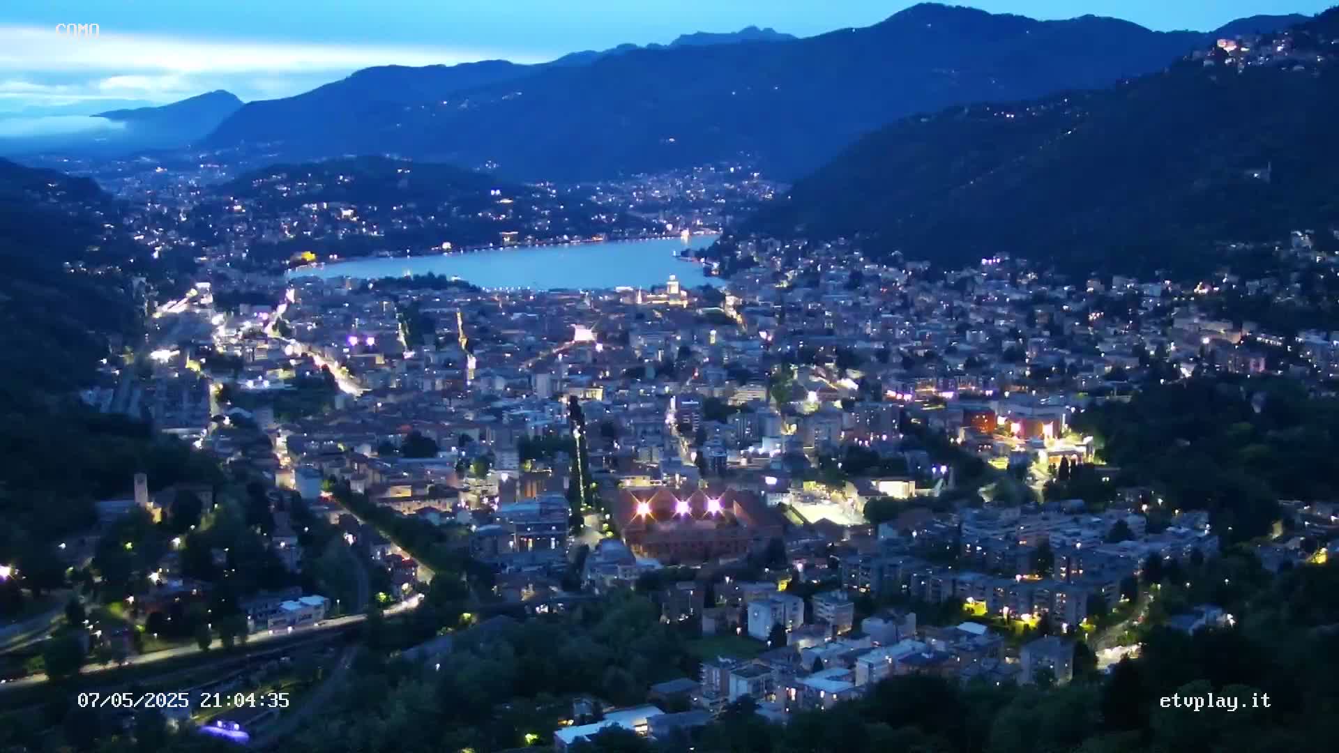 A nighttime, high-angle view shows a city nestled between a lake and mountains under a twilight sky.