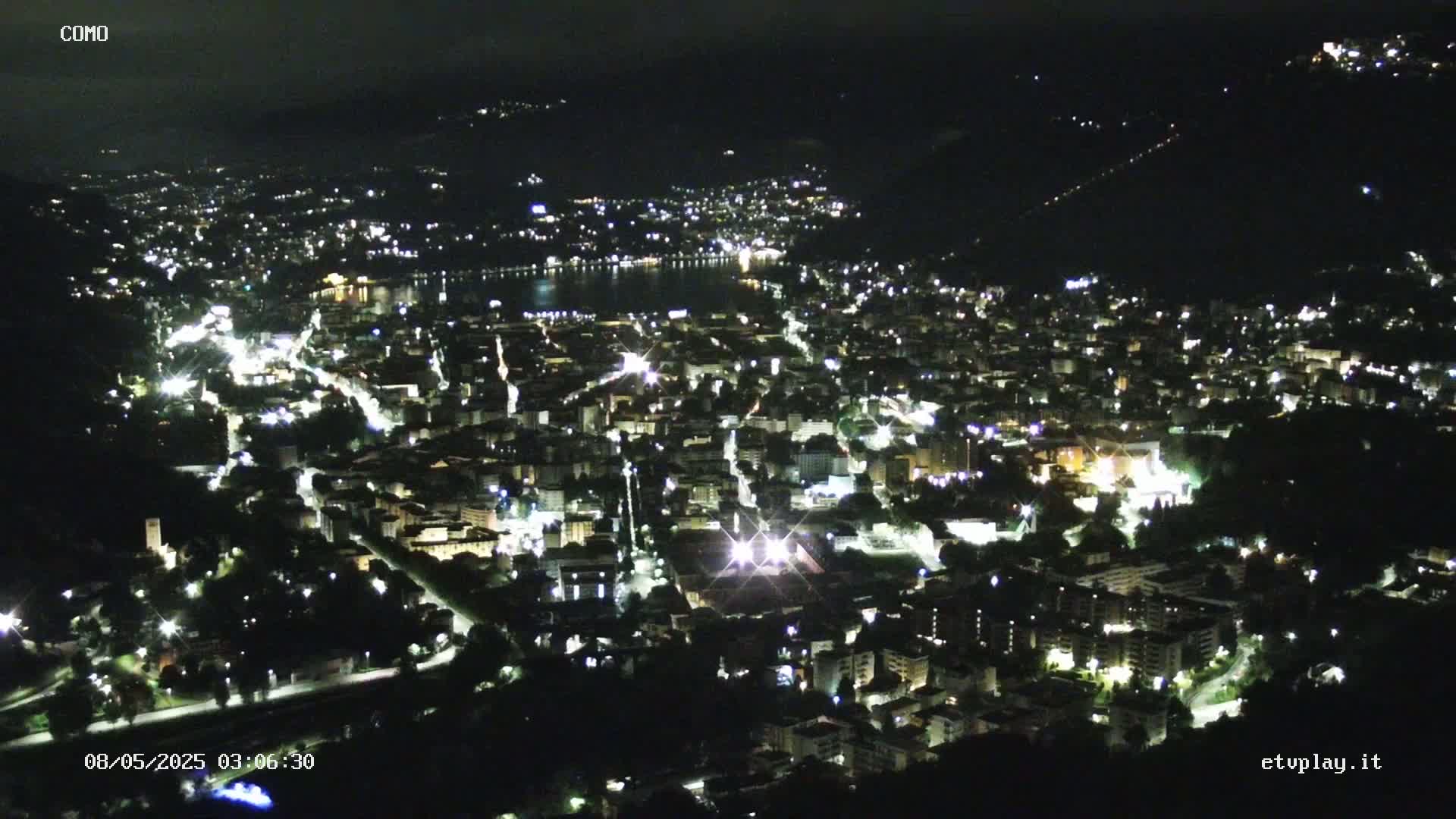 A nighttime aerial view shows a city illuminated by lights, nestled beside a dark body of water and surrounded by dark hills.