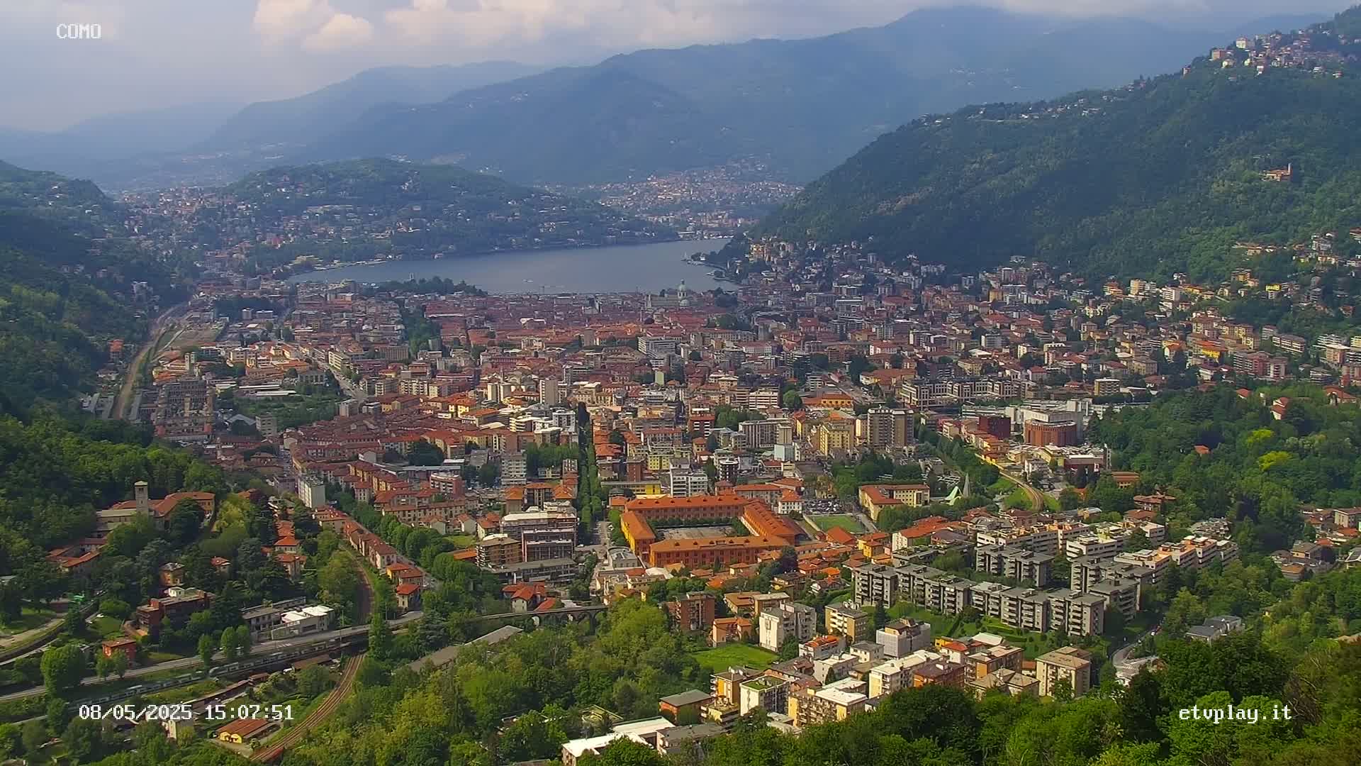 A sprawling city nestled in a valley between mountains, alongside a lake under a partly cloudy sky.