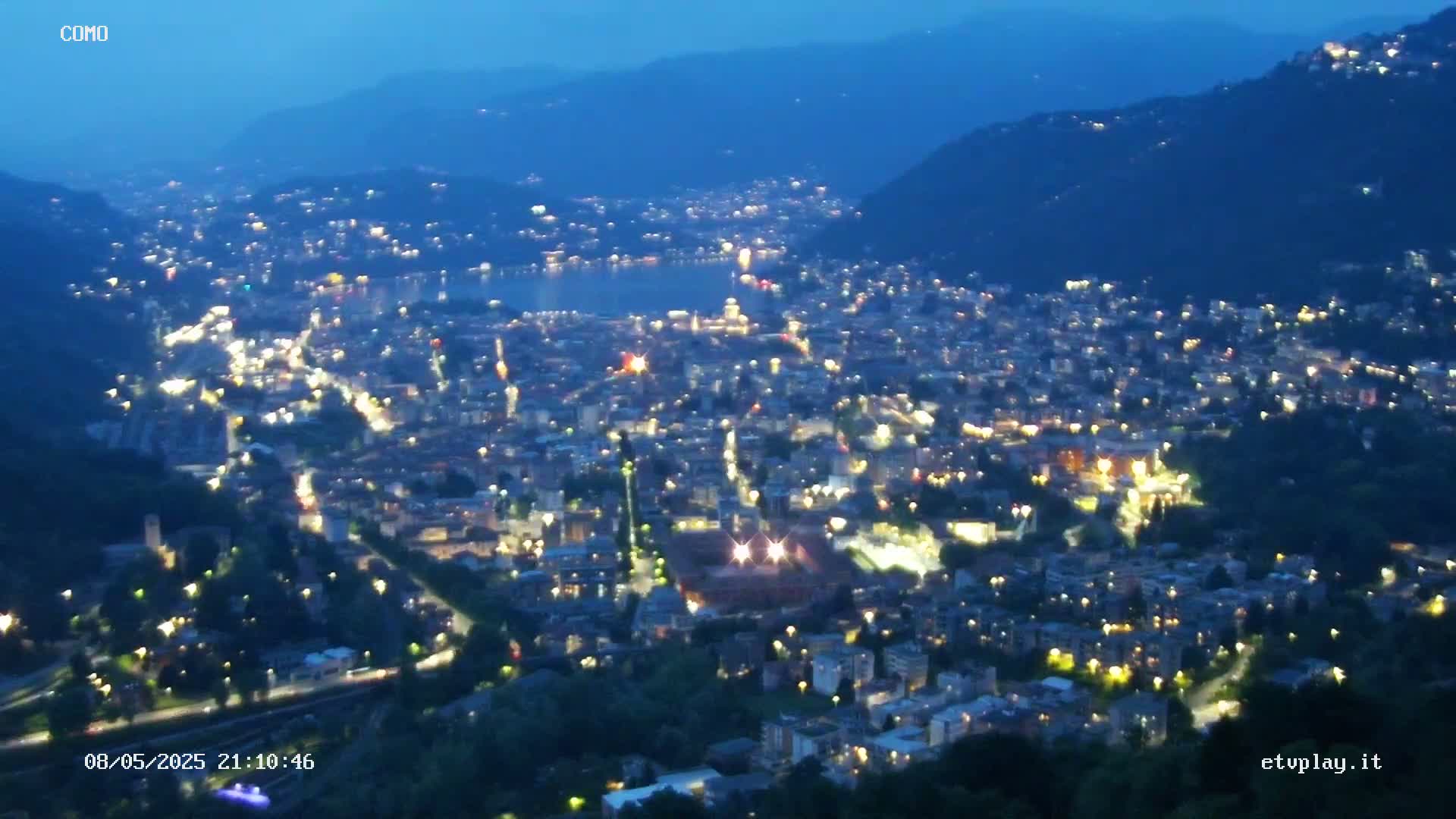 A nighttime view of a city nestled in a valley, illuminated by numerous lights, overlooking a dark body of water under a twilight sky.