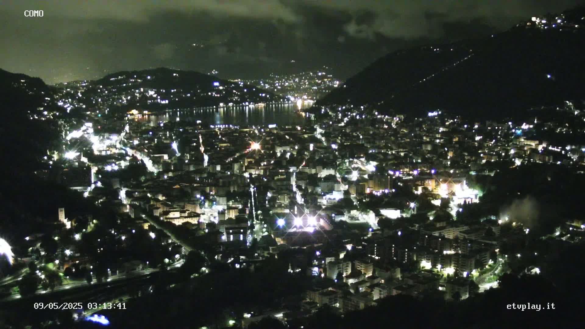 A nighttime aerial view of a city nestled between hills and a lake under a partly cloudy sky.