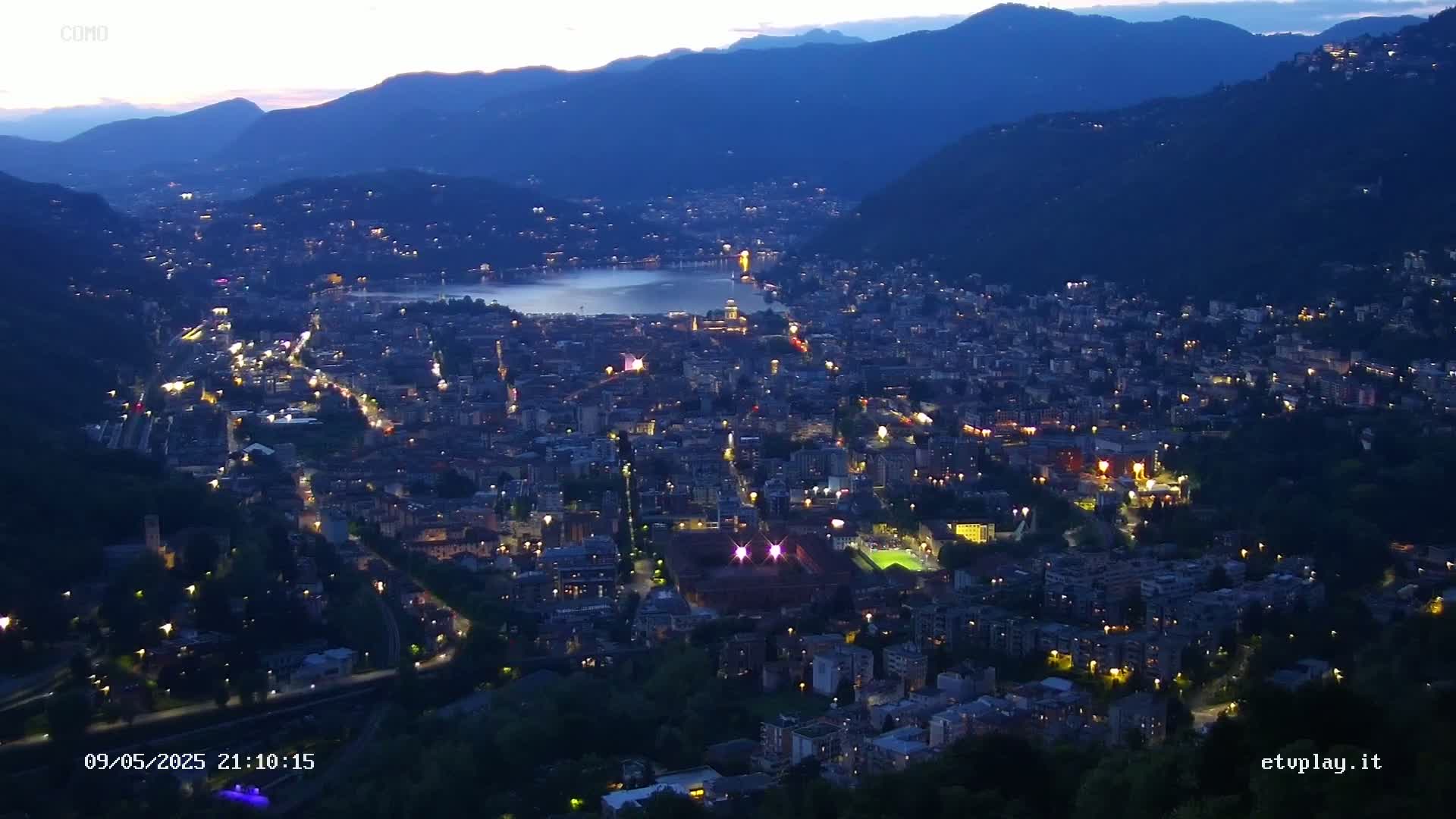 A nighttime, high-angle view of a city nestled in a valley between mountains, with lights illuminating the buildings and streets, and a body of water visible in the distance.