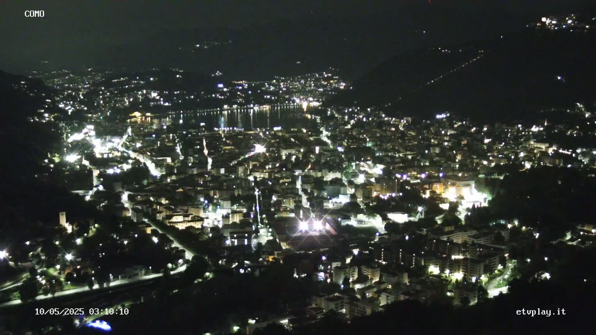 A nighttime aerial view shows a city illuminated by lights, nestled beside a body of dark water under a clear, dark sky.