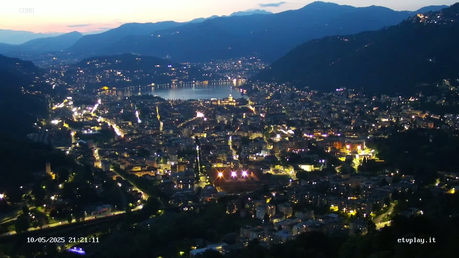 A nighttime aerial view of a city nestled in a valley beside a lake, under a twilight sky.