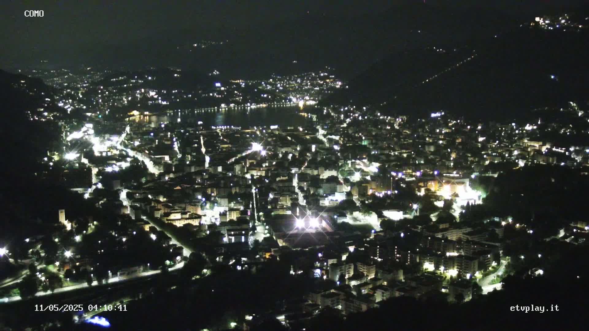 A nighttime, high-angle view shows a city illuminated by lights, nestled beside a dark body of water under a clear, dark sky.