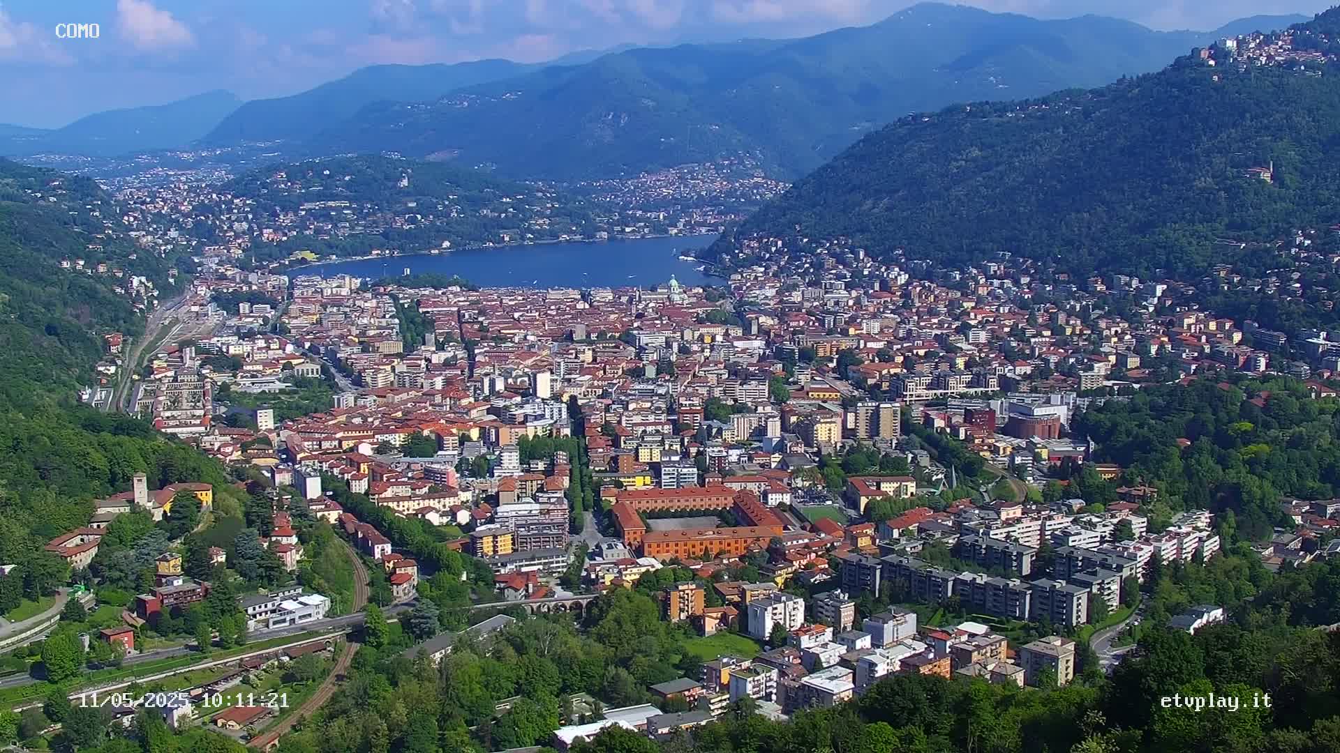 An aerial view of a city nestled in a valley between mountains, overlooking a lake on a sunny day.