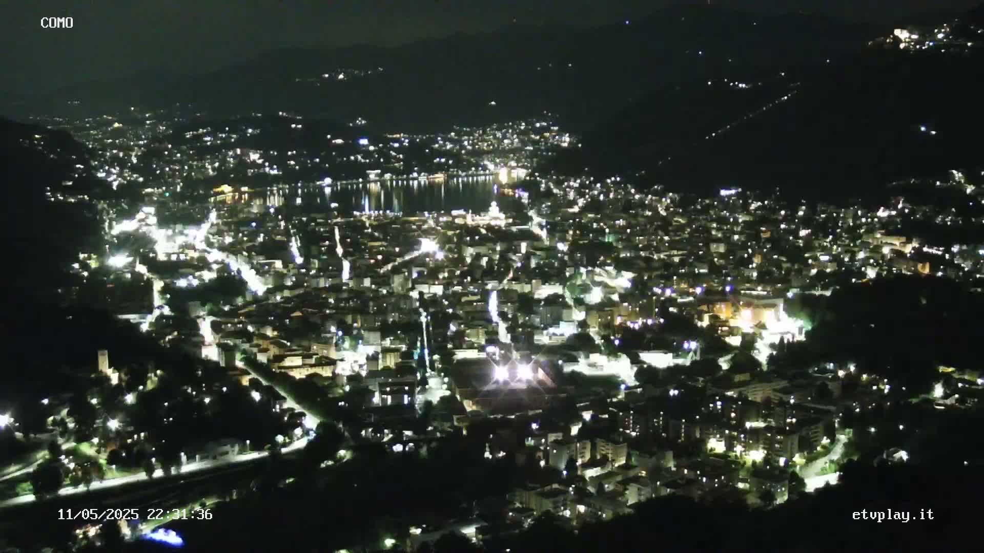 A nighttime, high-angle view shows a city illuminated by streetlights, nestled beside a dark body of water, with mountains in the background under a clear night sky.