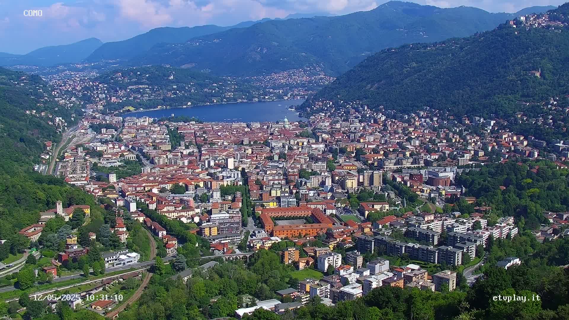 An aerial view shows a sprawling city nestled in a valley between mountains, overlooking a lake on a clear, sunny day.