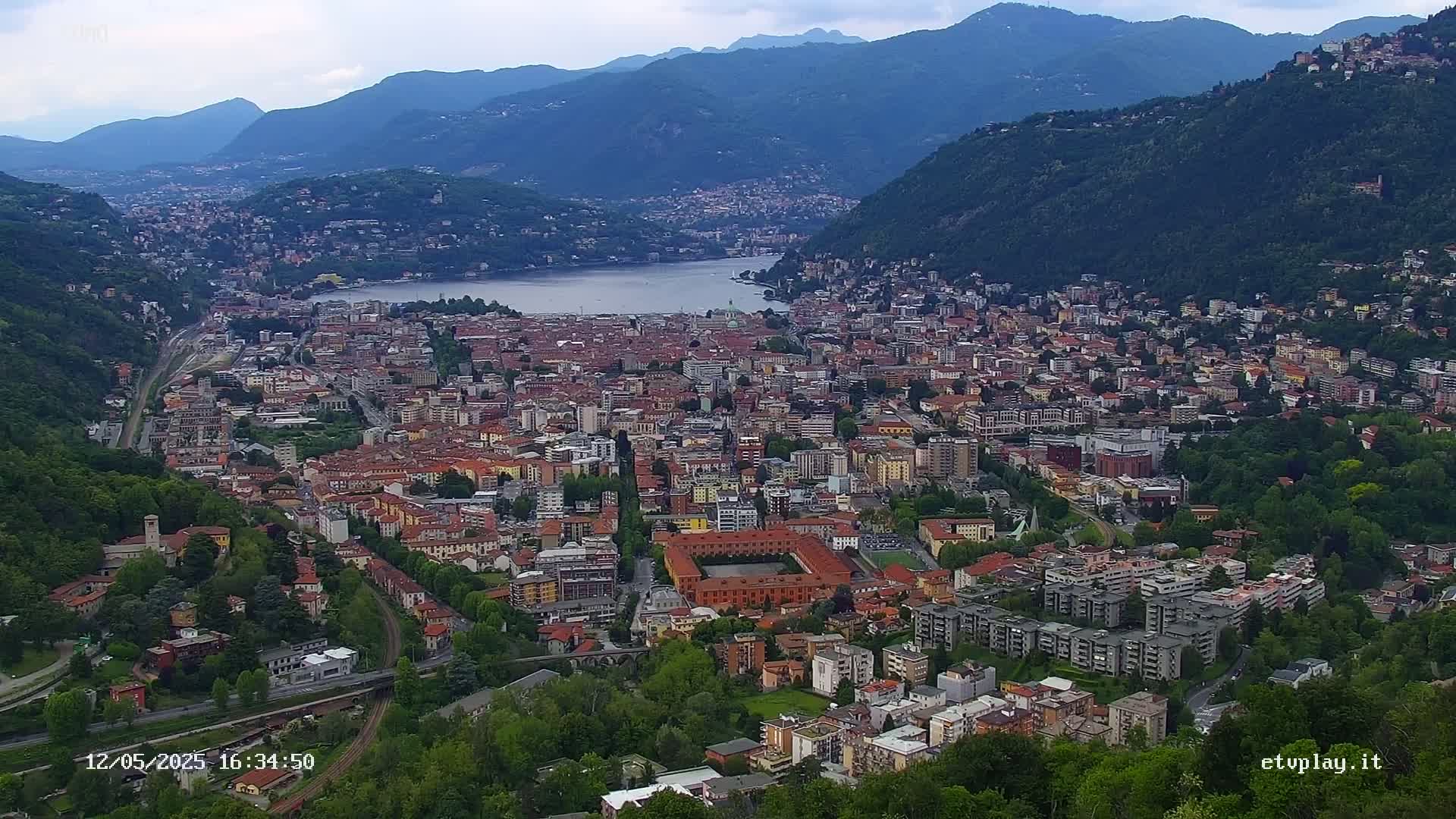 A sprawling city nestled between mountains and a lake, under a mostly cloudy sky.