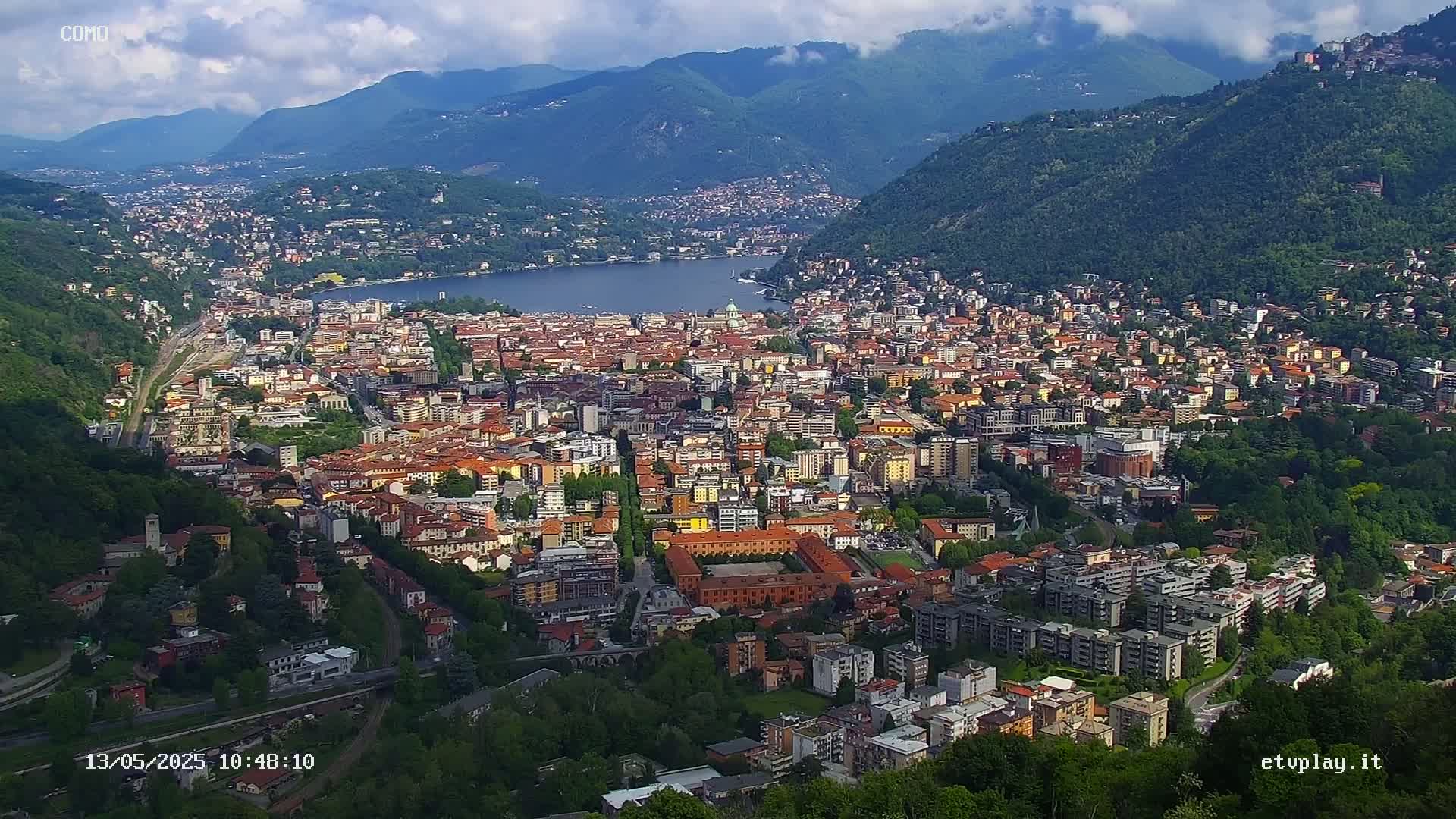 An aerial view shows a sprawling city nestled between a lake and mountains under partly cloudy skies.