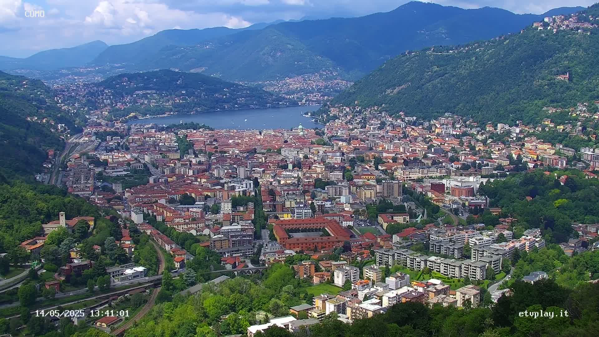 A sprawling city nestled in a valley between mountains, overlooking a lake under partly cloudy skies.