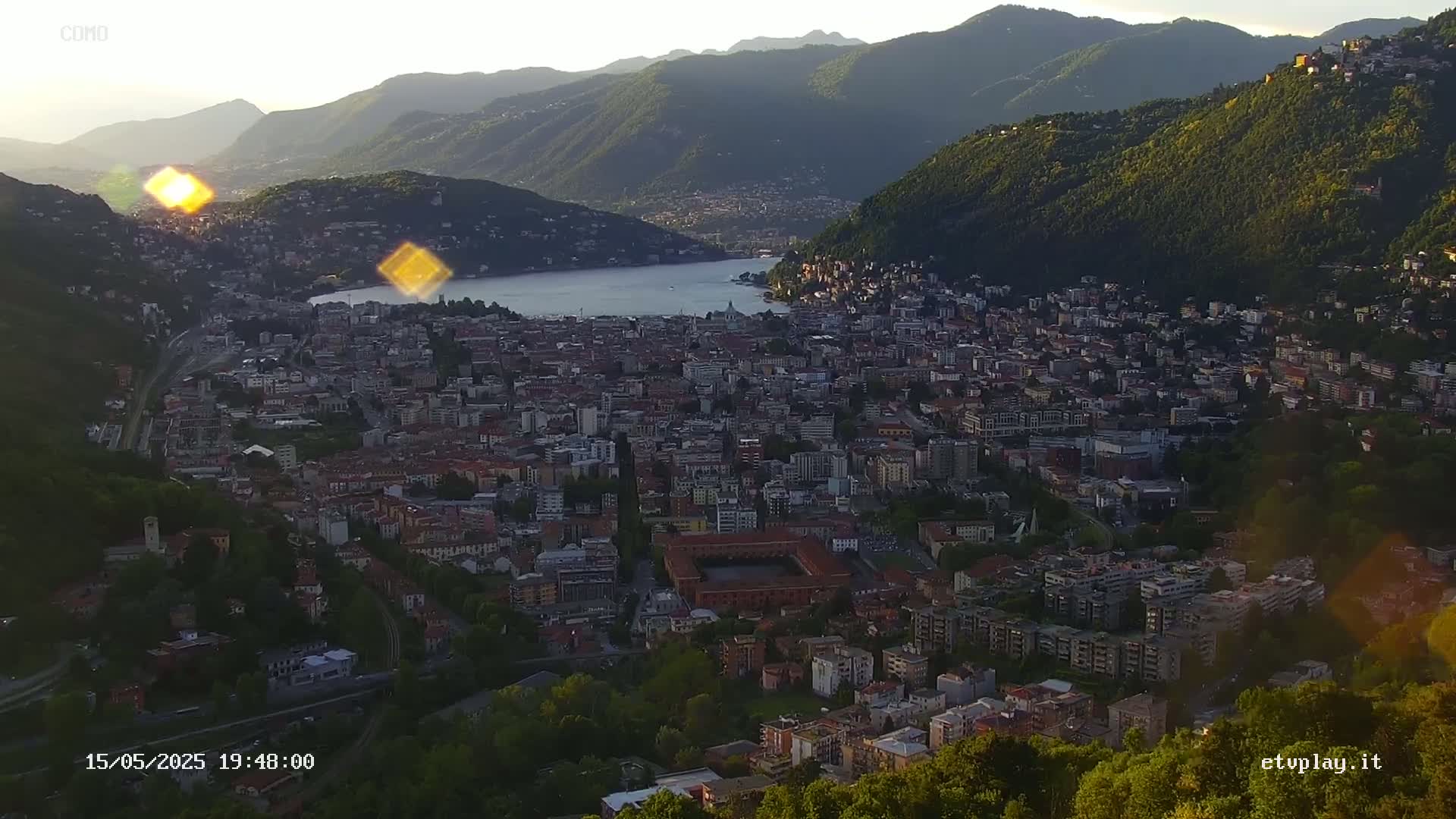 A sprawling city nestled between mountains and a lake at sunset, with the sun creating lens flares.