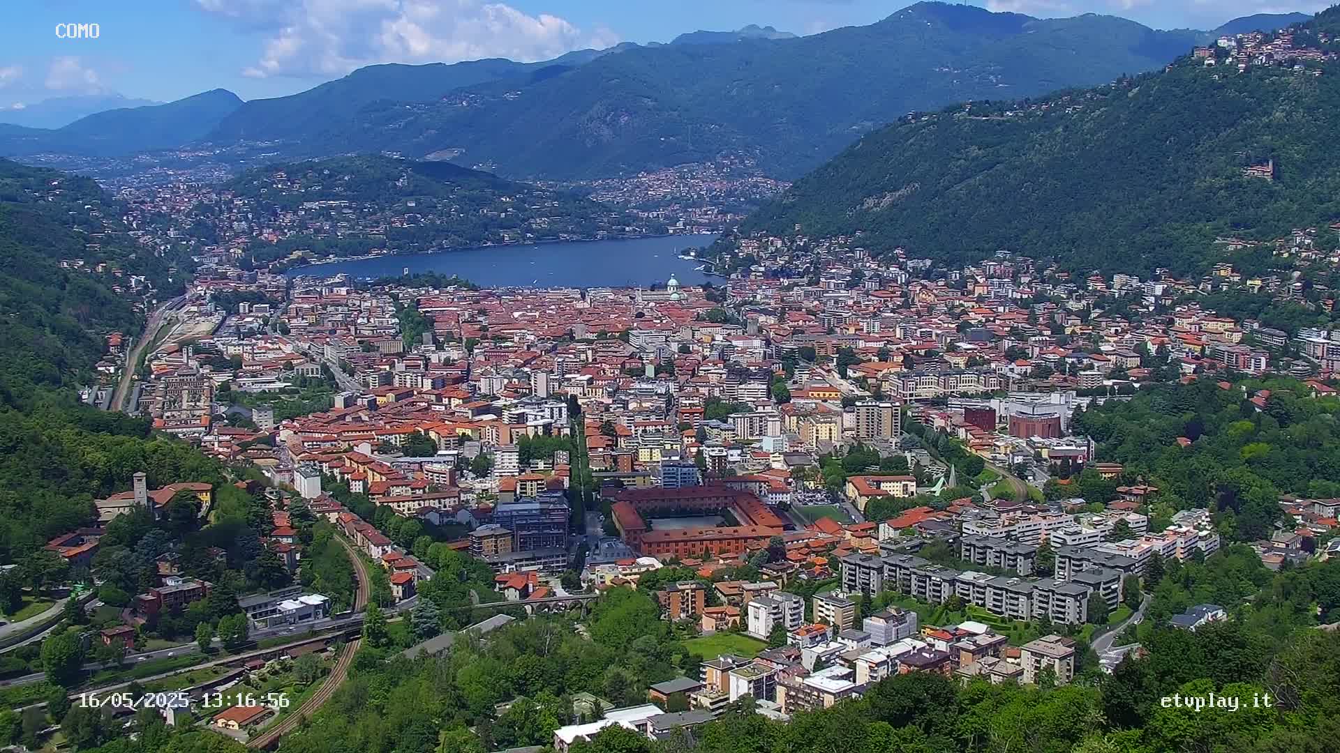 A sprawling city nestled in a valley between mountains, overlooking a lake on a partly sunny day.