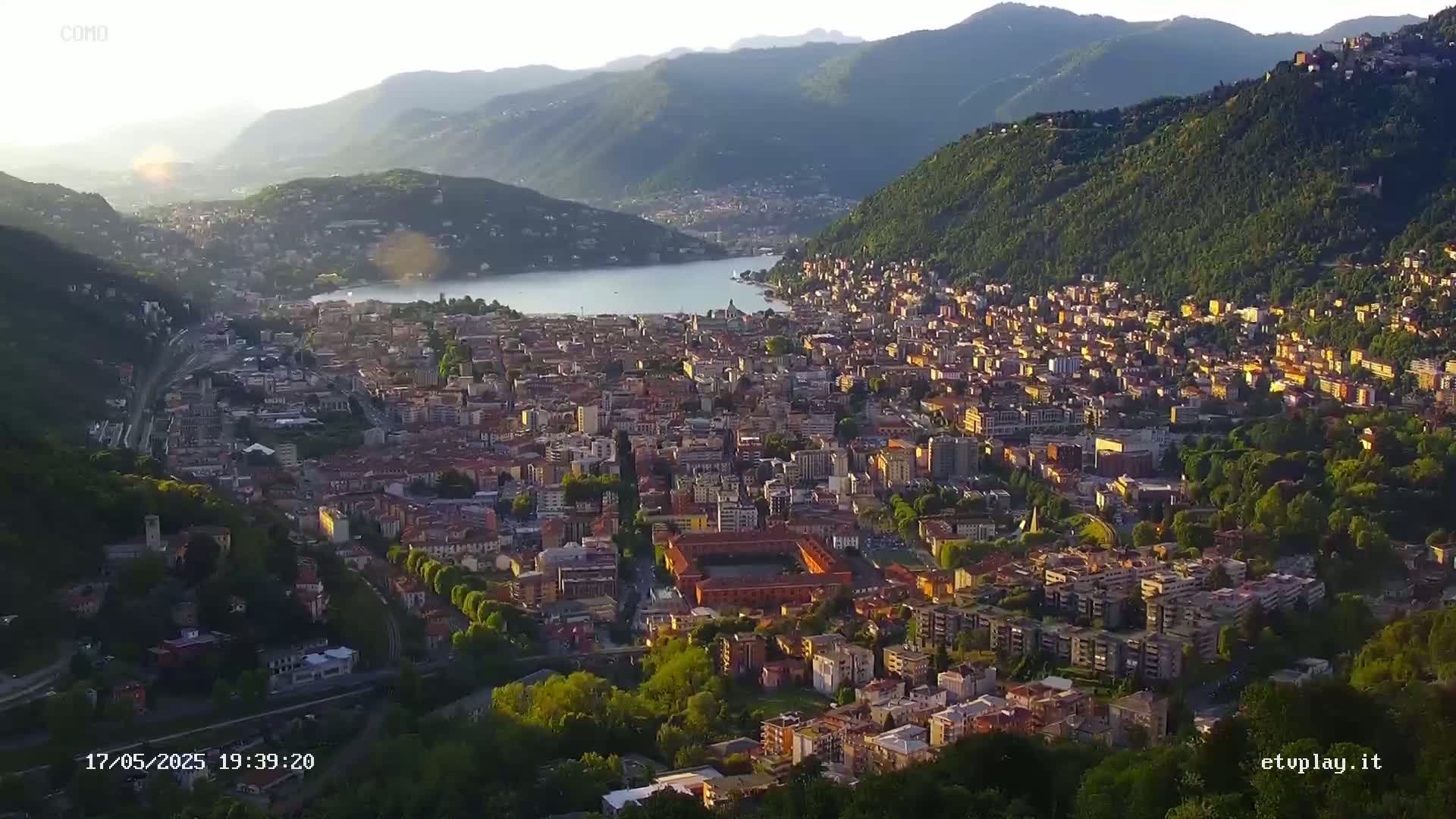 An aerial view shows a city nestled between mountains and a lake at sunset, under a clear sky.