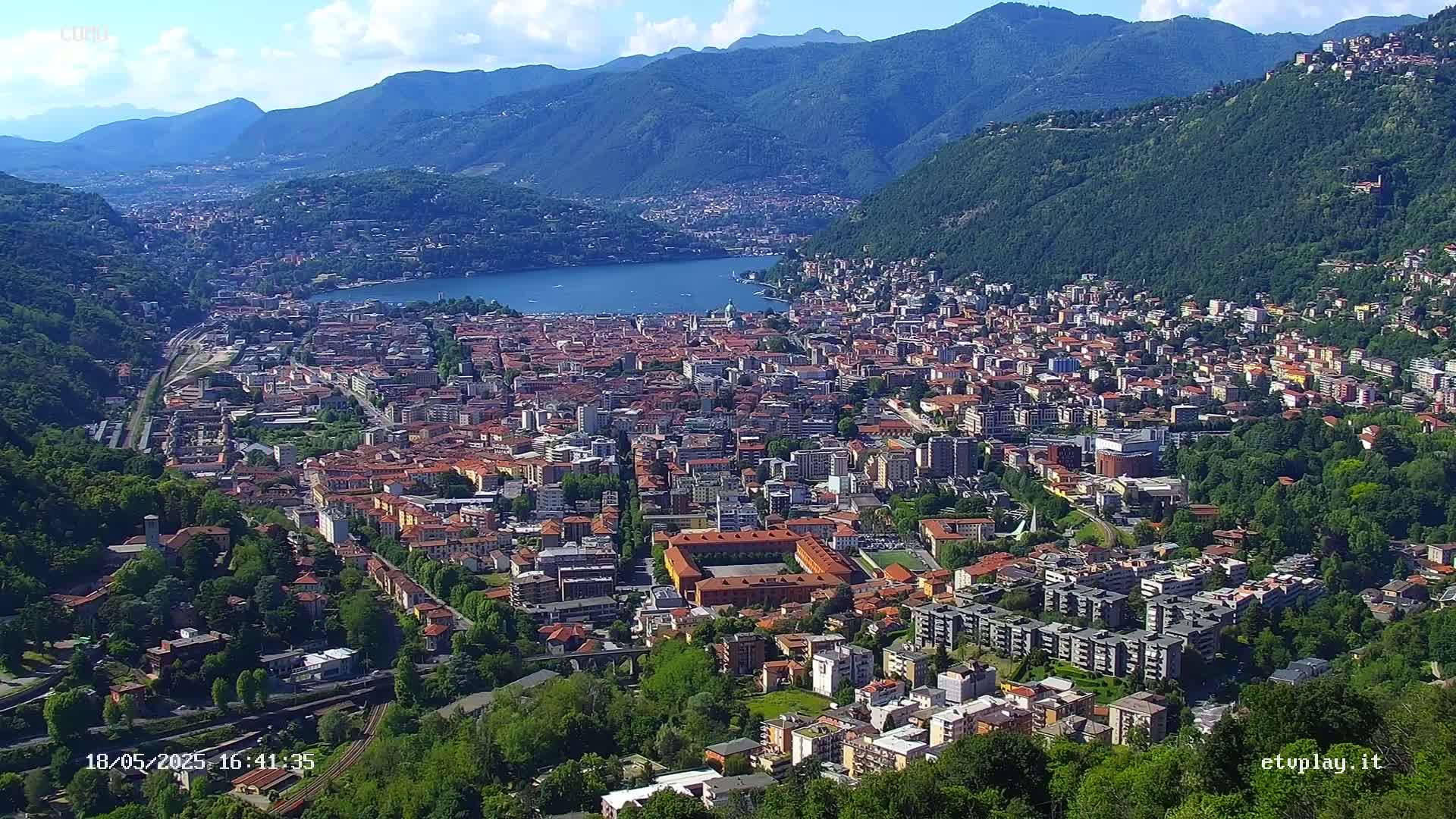 An aerial view shows a city nestled in a valley between mountains on a sunny day, with a lake visible in the distance.