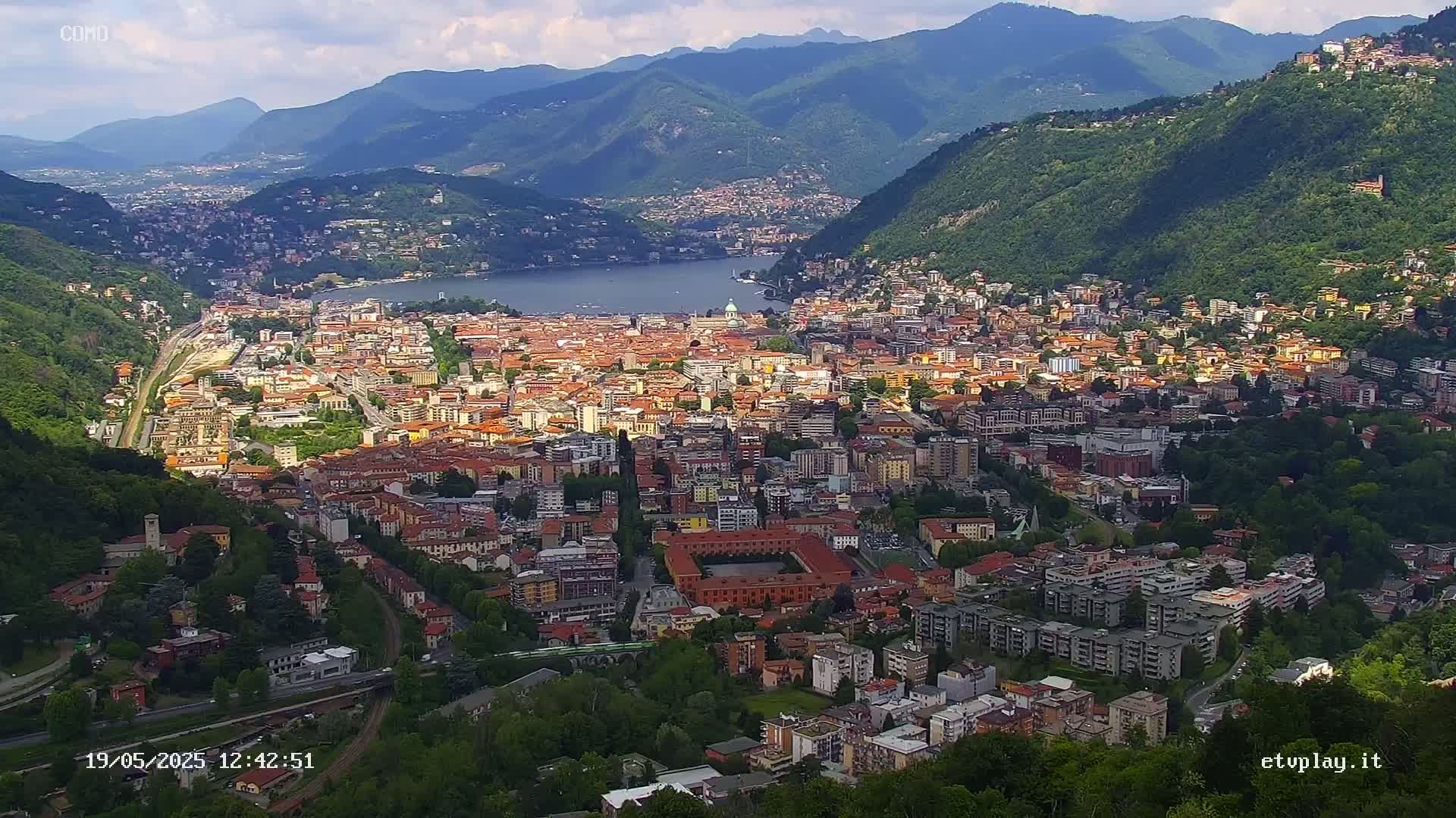 An expansive city nestled in a valley between mountains, overlooking a lake under partly cloudy skies.