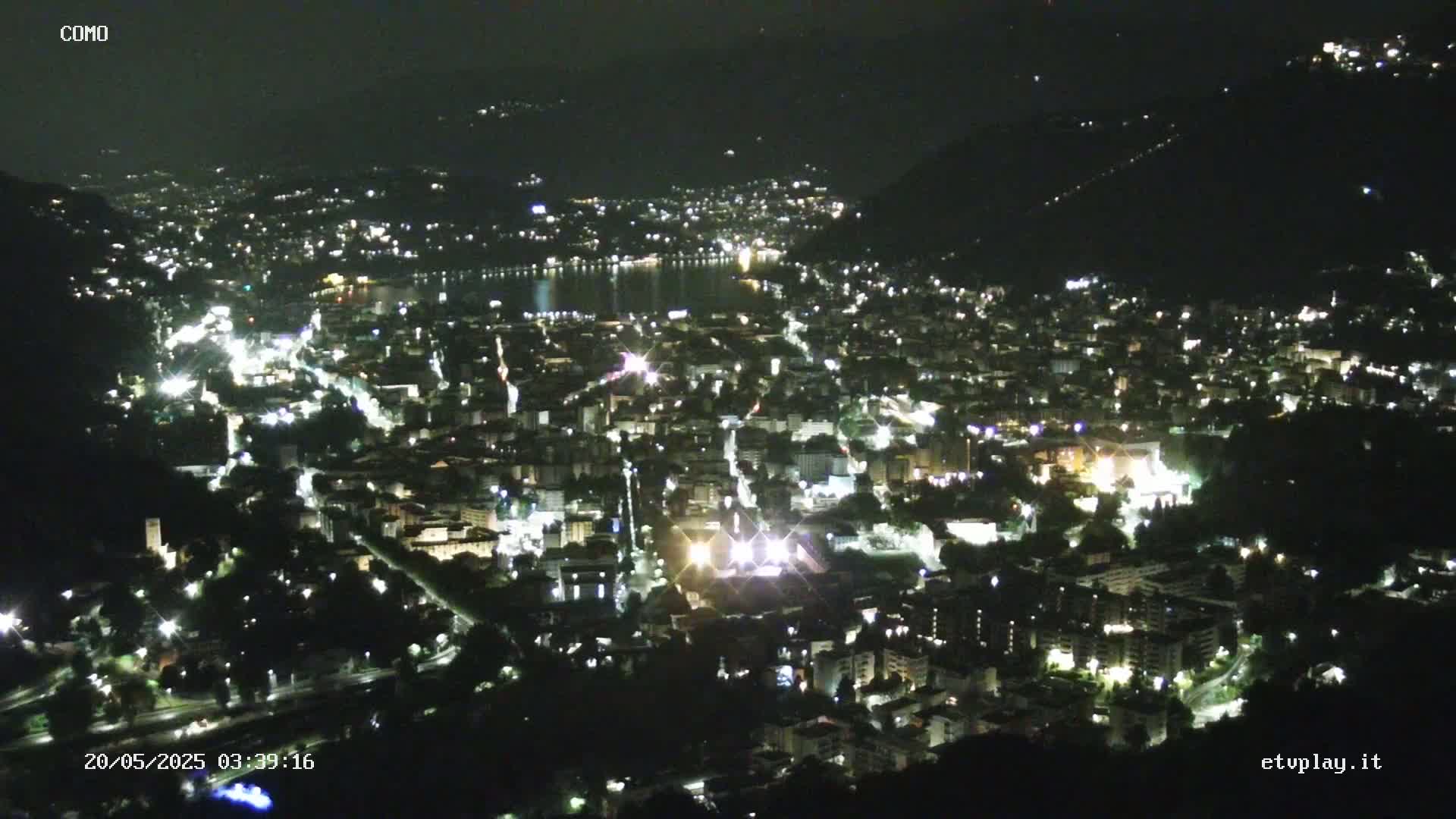 A nighttime, high-angle view shows a city nestled beside a body of water, illuminated by numerous lights.