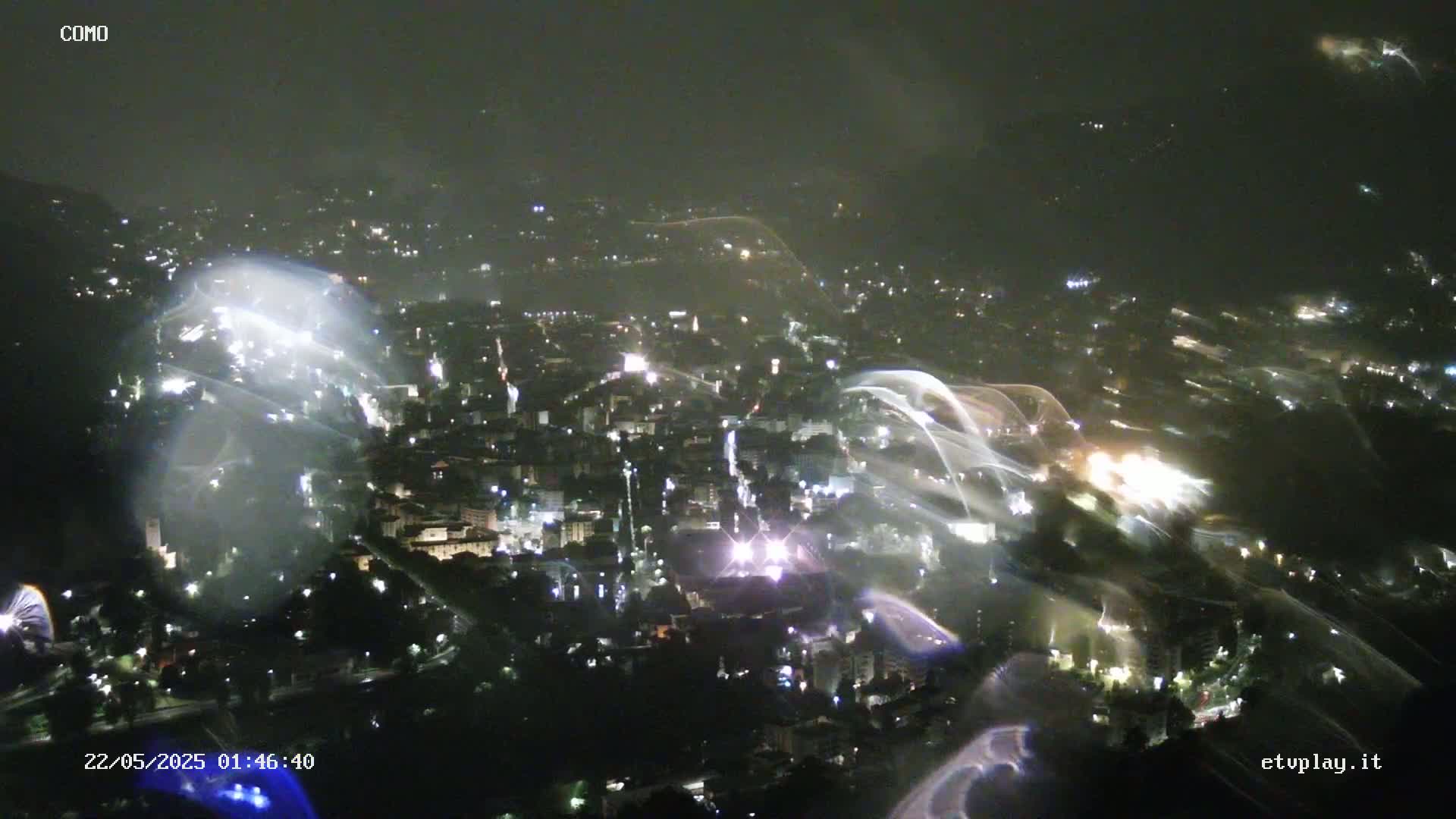 A nighttime, high-angle view of a city illuminated by lights, with streaks of light possibly from rain or camera malfunction in a hazy atmosphere.