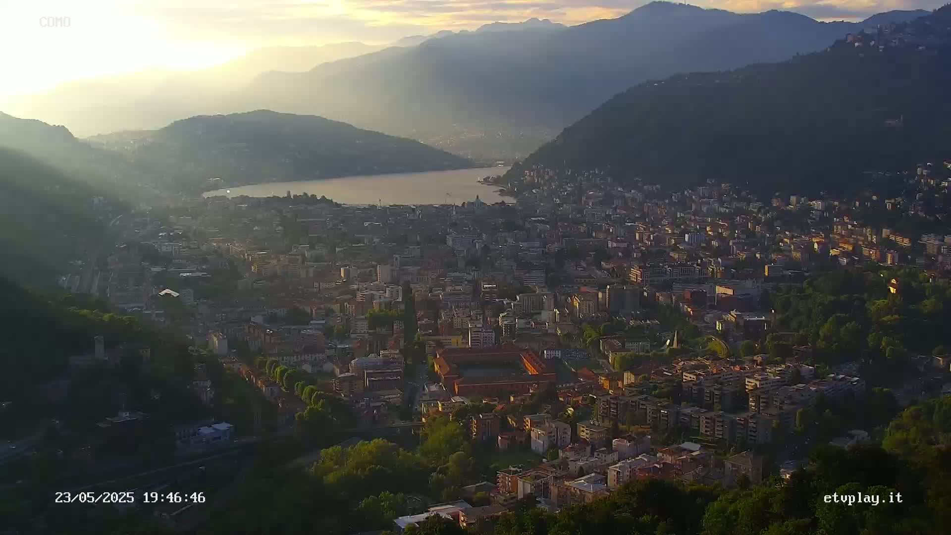 A hazy sunrise illuminates a sprawling city nestled between a lake and mountains.
