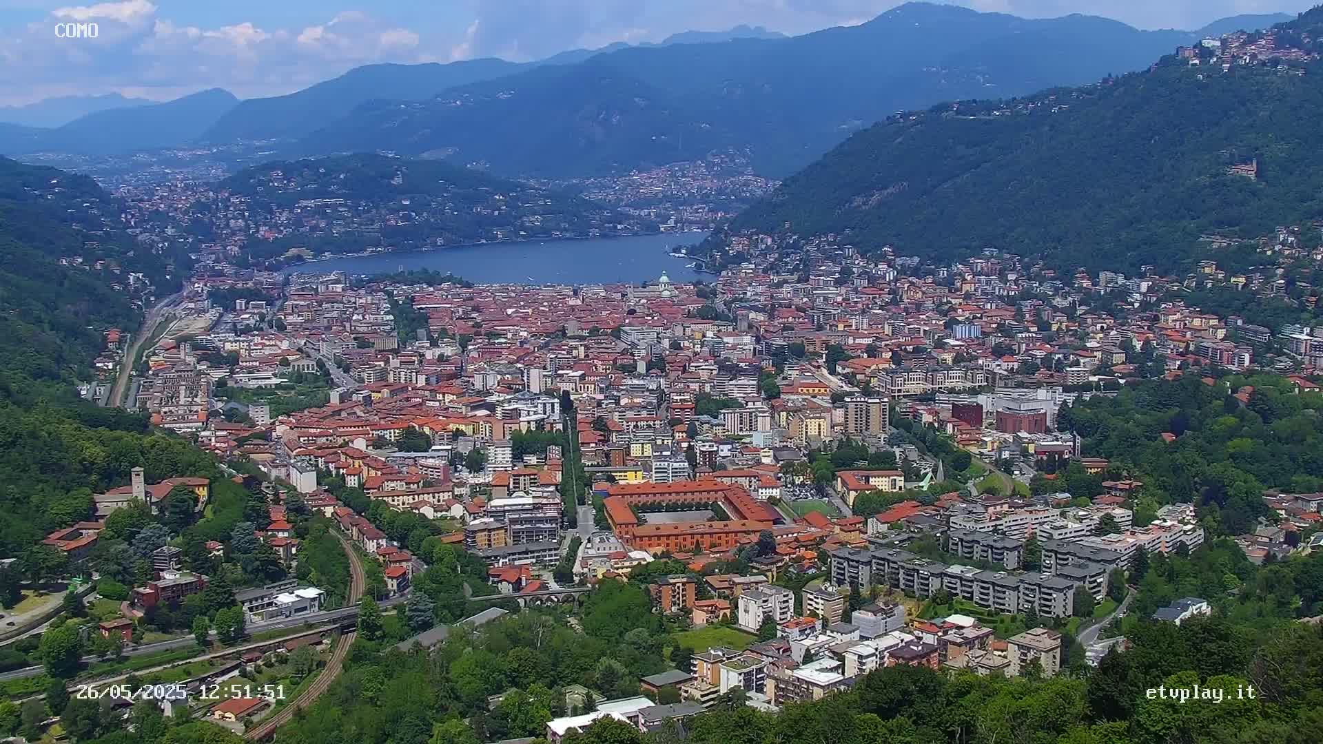 An aerial view shows a city nestled in a valley between mountains, next to a lake on a clear, sunny day.