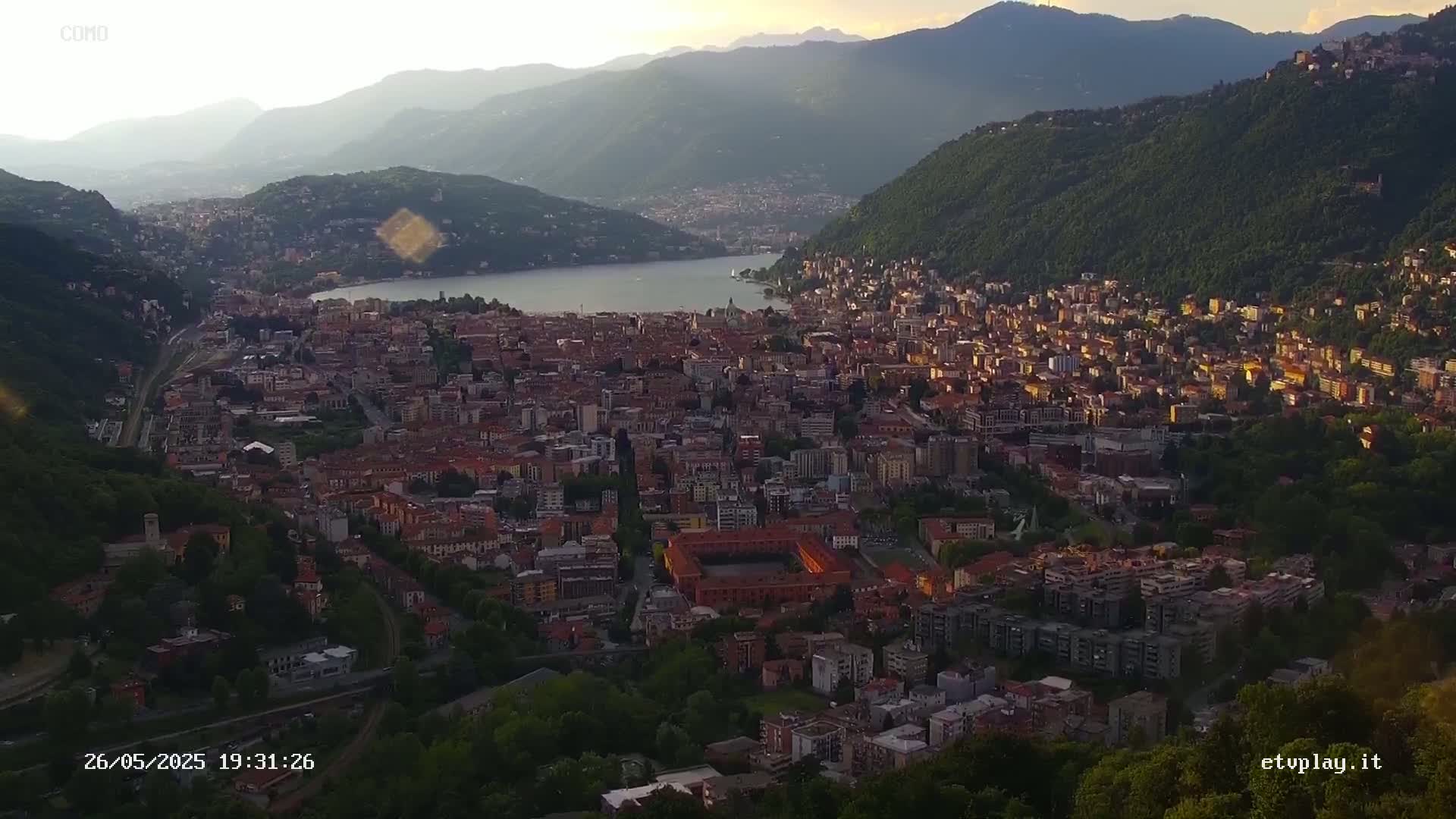 An aerial view shows a large city nestled in a valley between mountains, next to a lake, at sunset.
