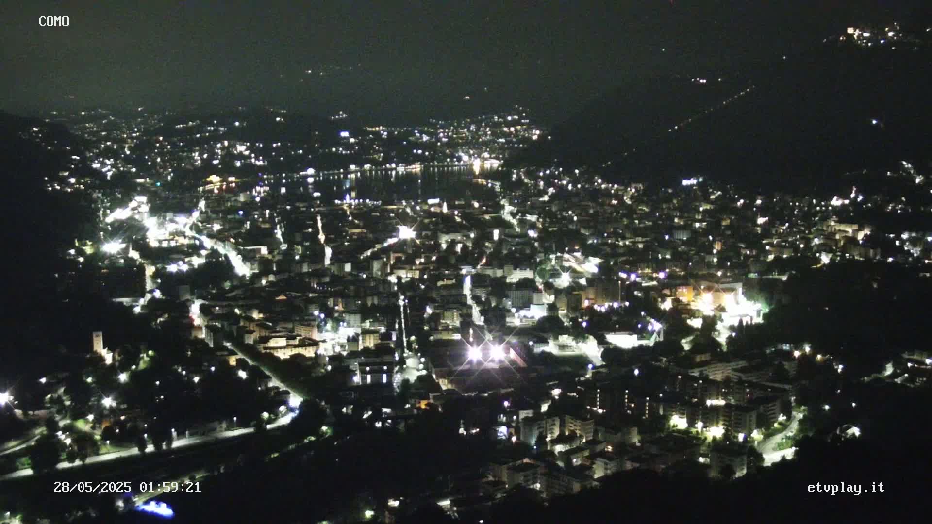 A nighttime aerial view of a city illuminated by lights, situated near a body of water under a clear, dark sky.